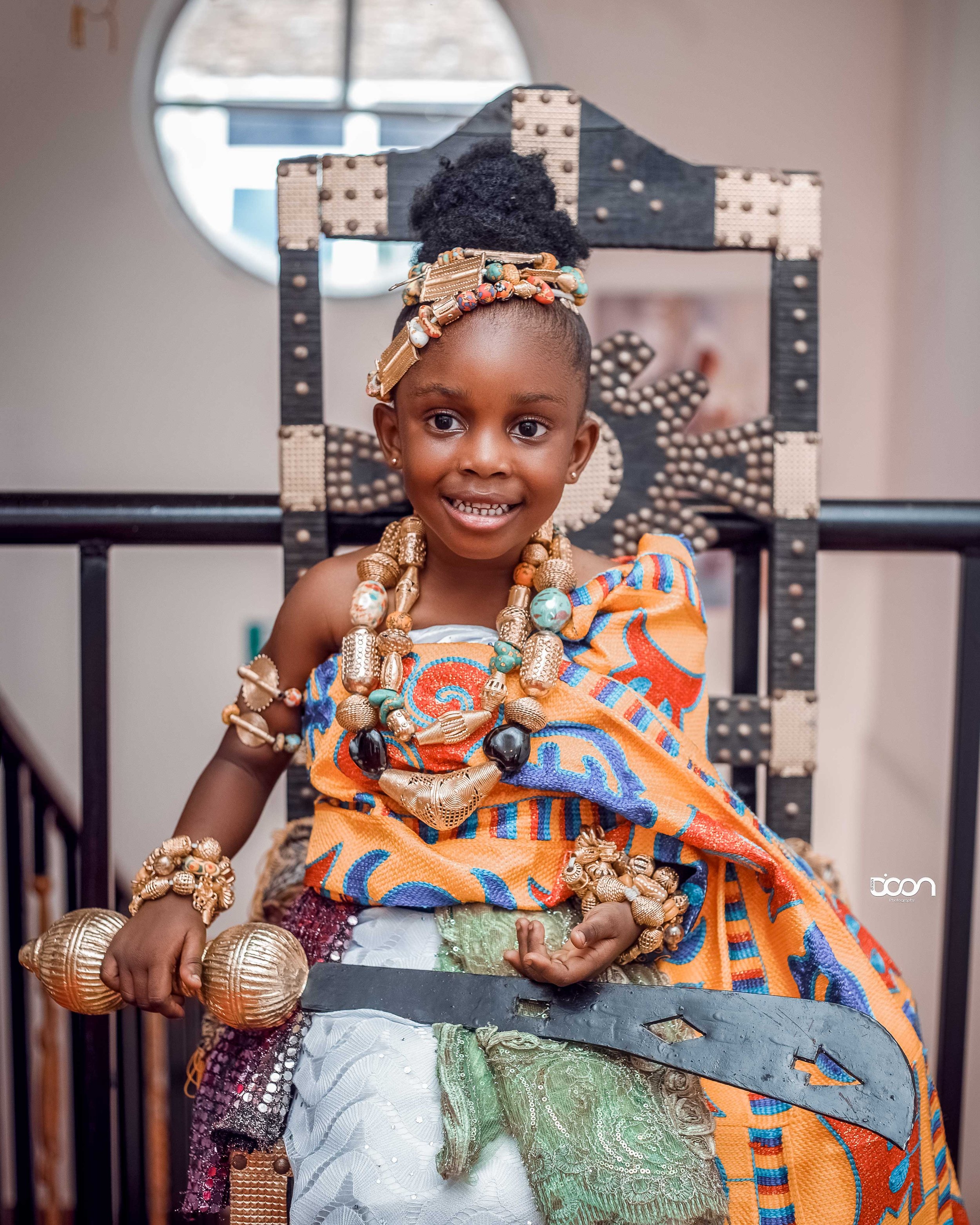 Young girl dressed in vibrant traditional African attire, sitting on an ornate wooden throne, adorned with large gold jewelry and holding a sword.