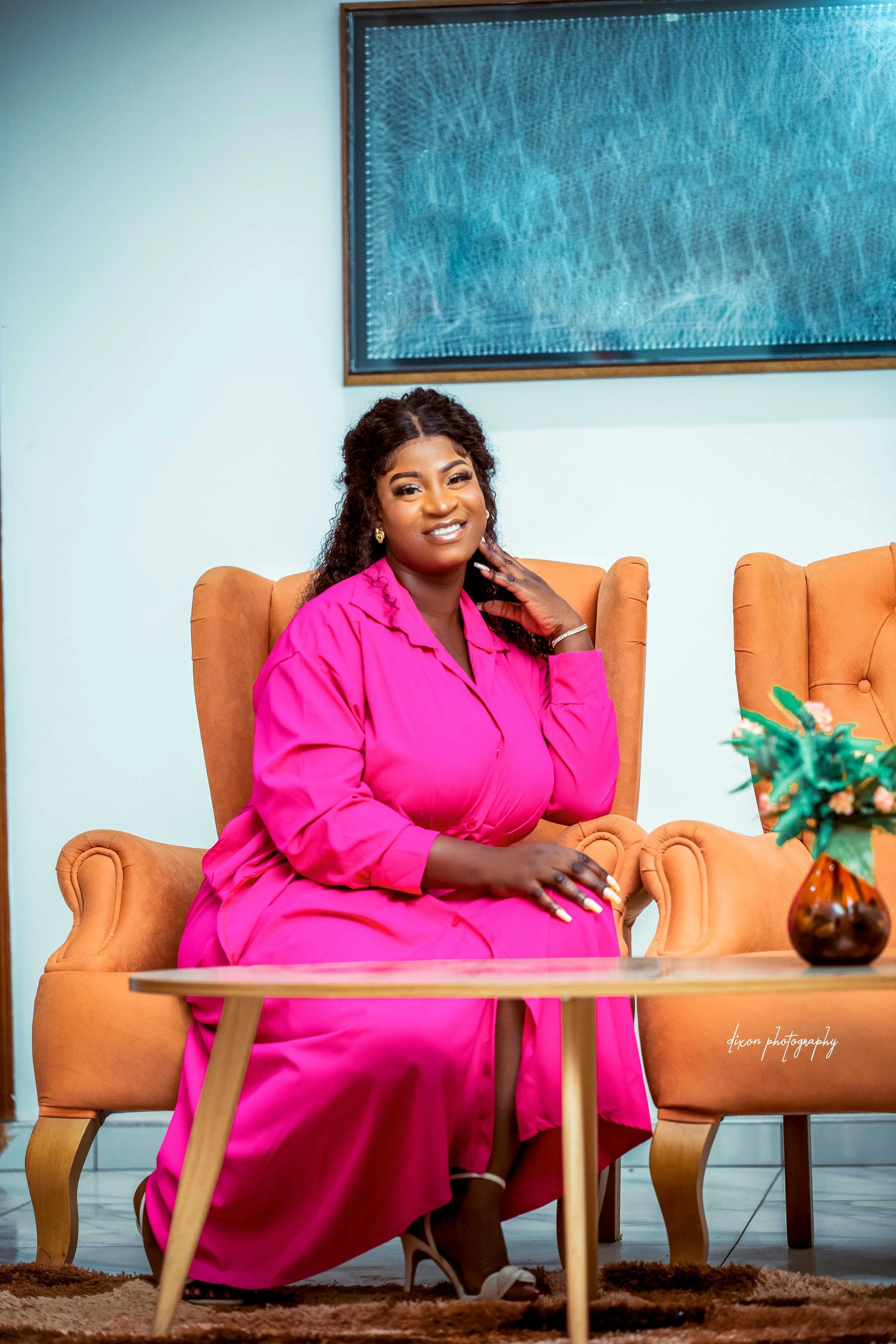 A woman with dark curly hair wearing a bright pink dress sits on a tan armchair in a room with a light-colored wall, a floor rug, and a wooden table with a flower vase. She is smiling and looking at the camera.