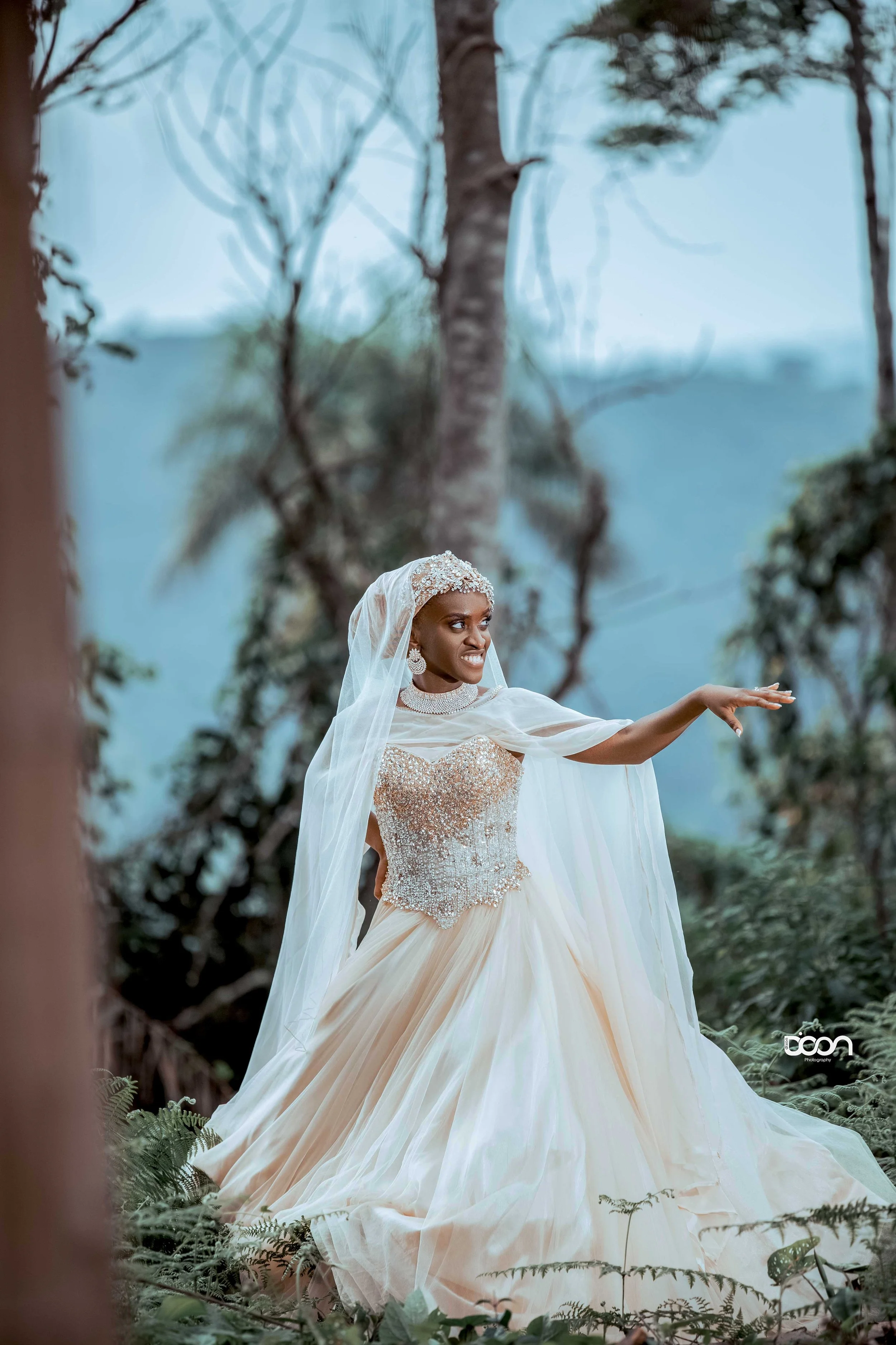A woman in a wedding dress stands outdoors in a forested area with trees and plants, smiling and reaching out with her right arm.