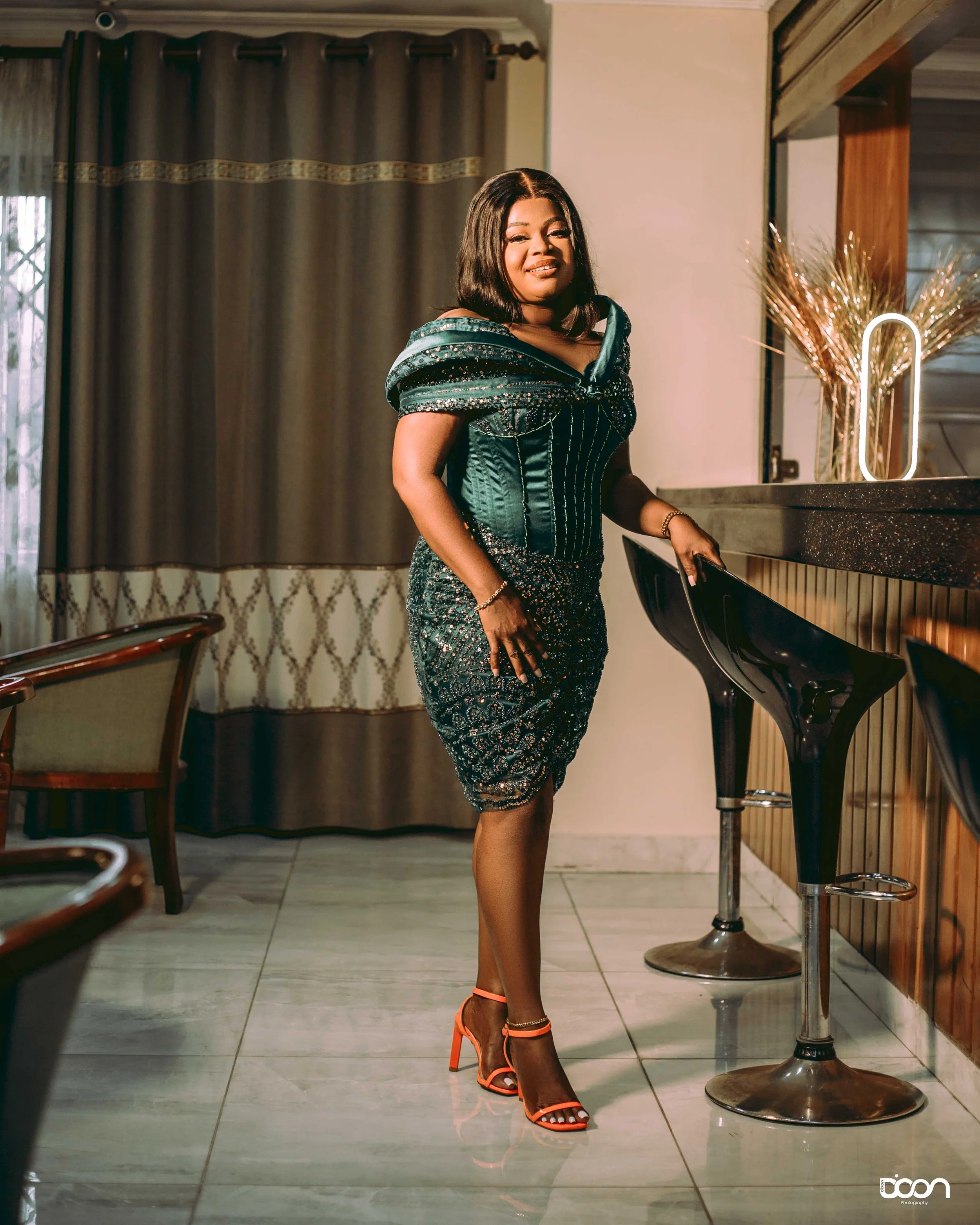 A woman in a dark teal, ornate, off-the-shoulder gown with intricate detailing, standing inside a room with a bar. She is wearing coral high heels, gold jewelry, and is smiling at the camera while resting her hand on the bar counter.
