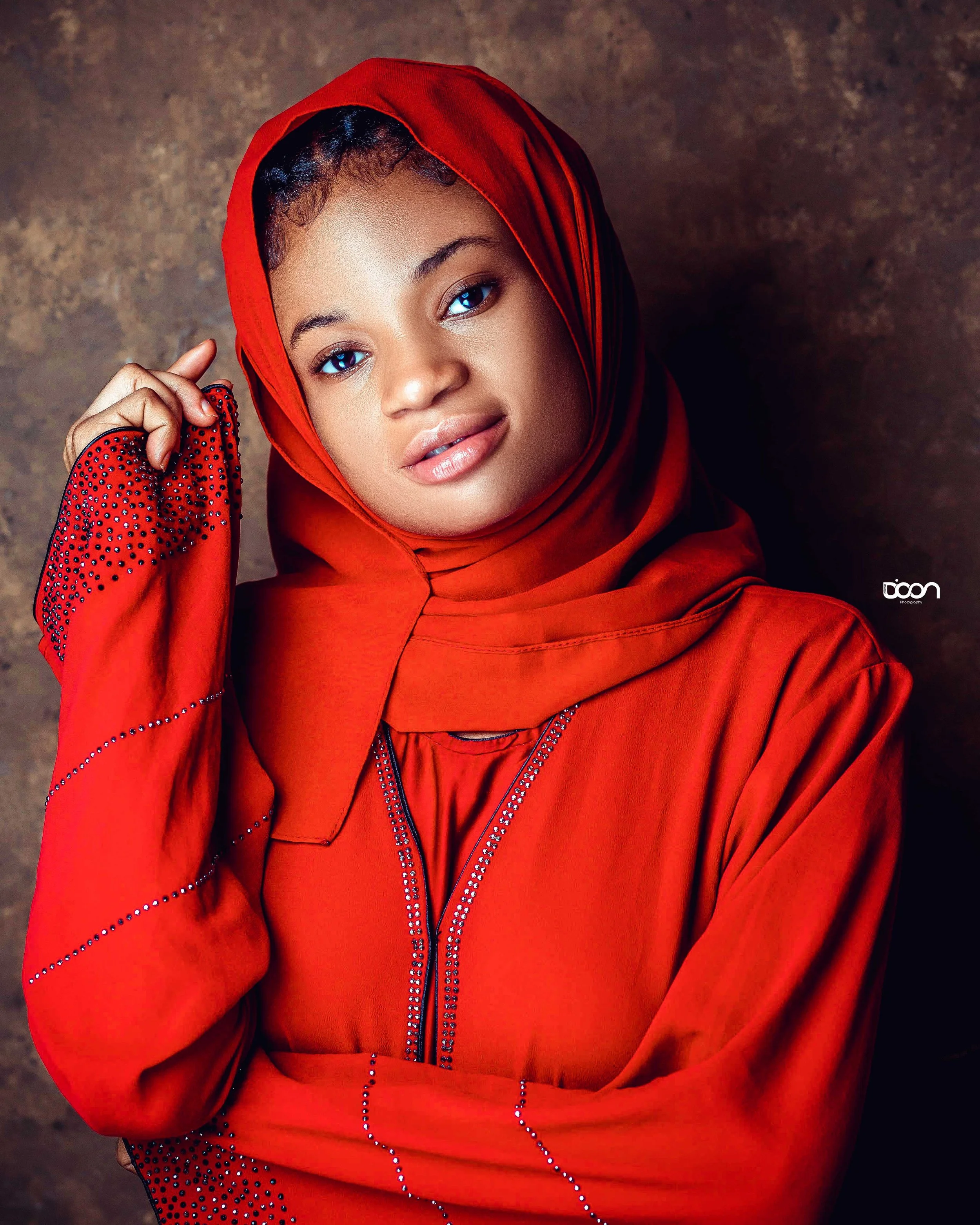 A woman with a medium-dark skin tone wearing a red hijab and matching red dress with rhinestone embellishments. She is posing with her hand near her head against a textured brown background.