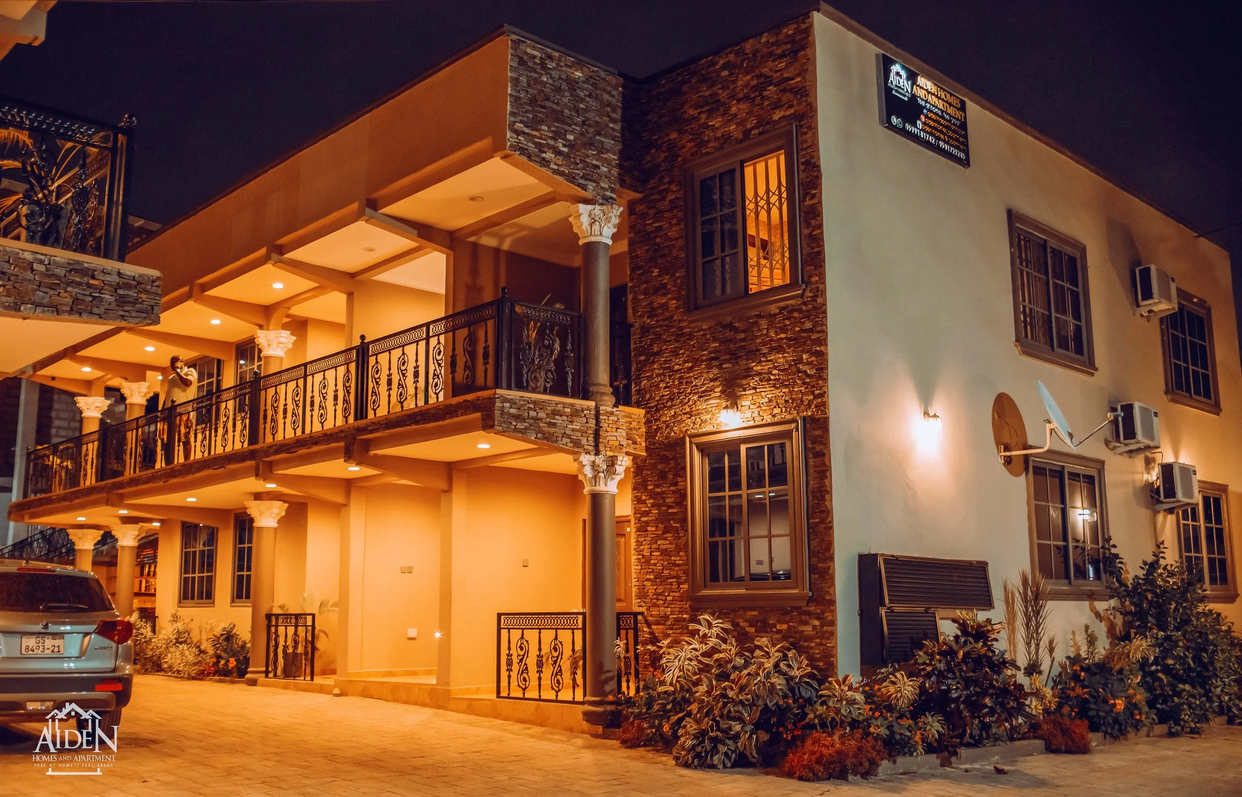 A modern multi-story building at night with warm lighting, multiple balconies with decorative railings, stone accents, and several windows with curtains. There are air conditioning units on the side, a satellite dish, and a parking area with a car.