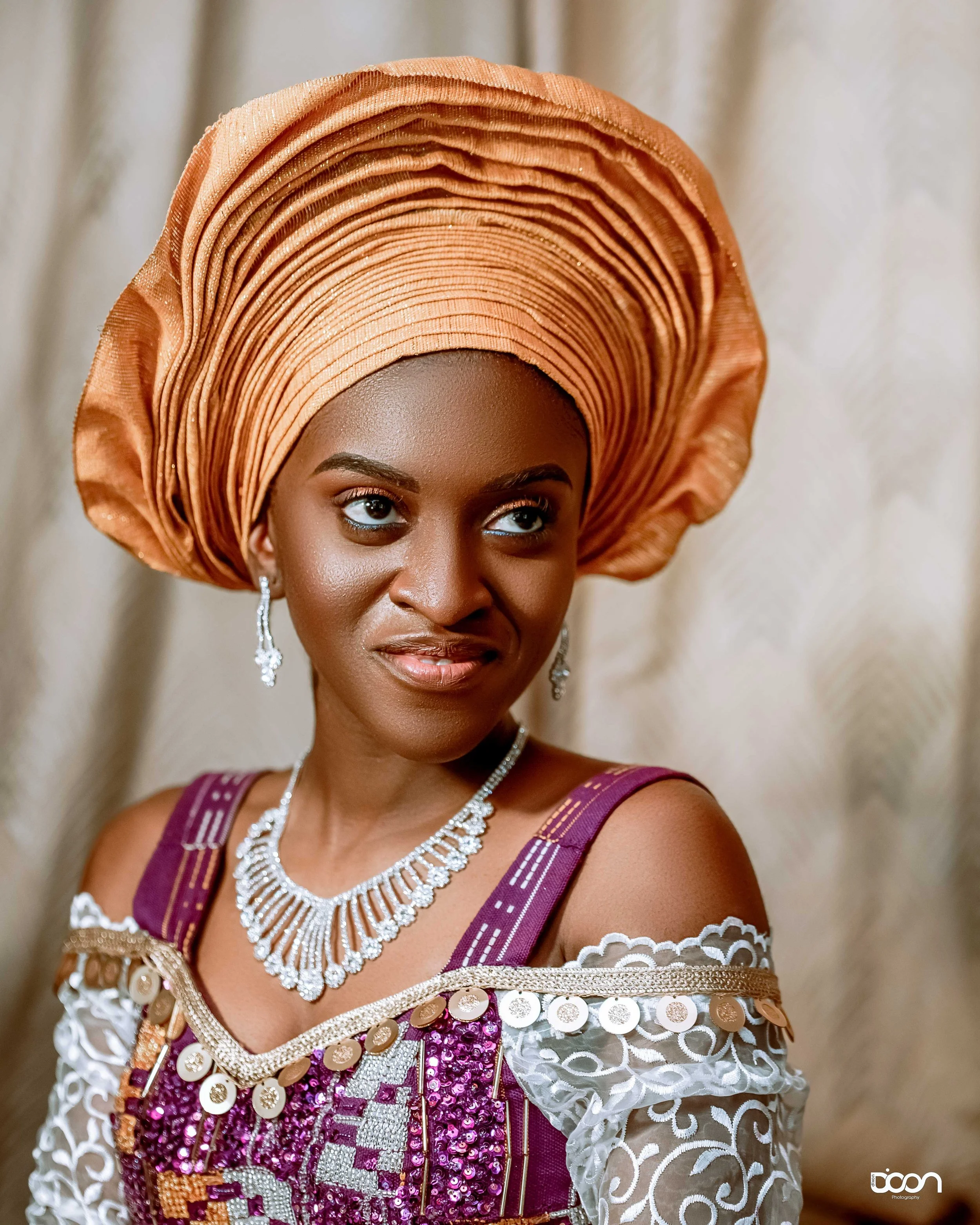 A woman wearing an elegant purple dress with intricate white lace and gold embellishments, topped with a large, peach-colored gele and adorned with sparkling jewelry, including earrings, a necklace, and a bracelet.