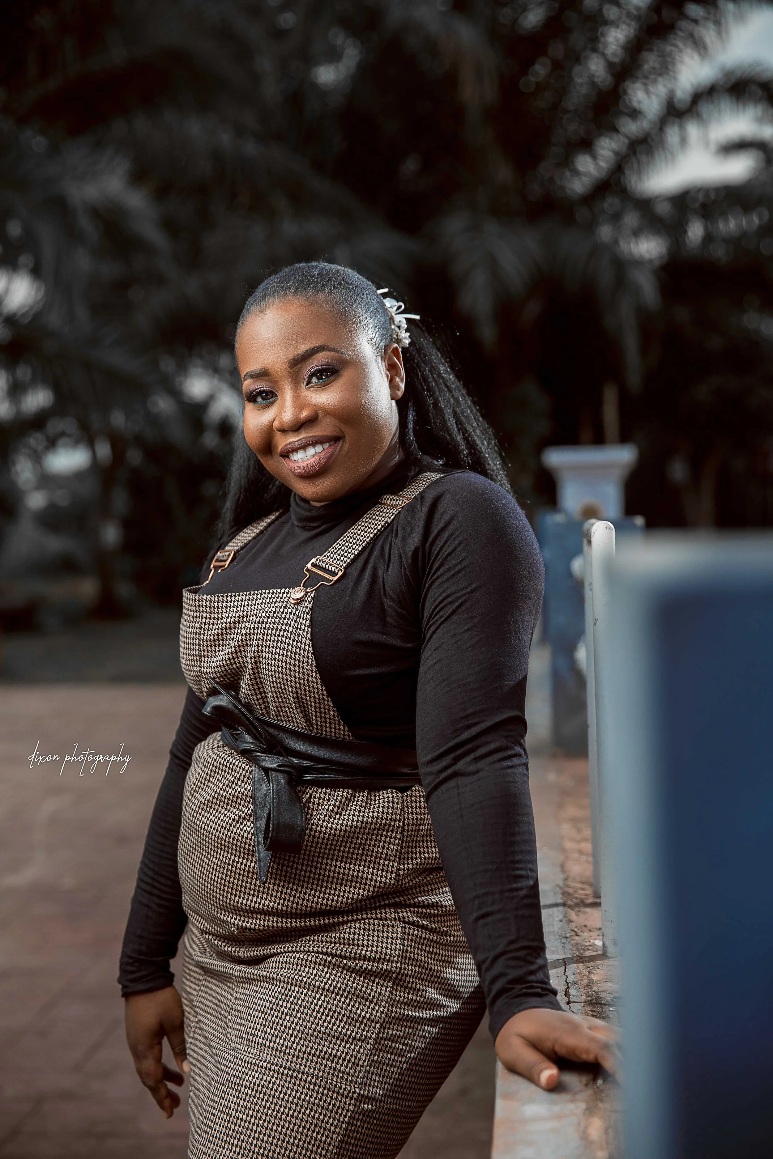 A smiling woman wearing a black long-sleeve top and beige checkered overalls, standing outdoors near a blue railing with greenery in the background.
