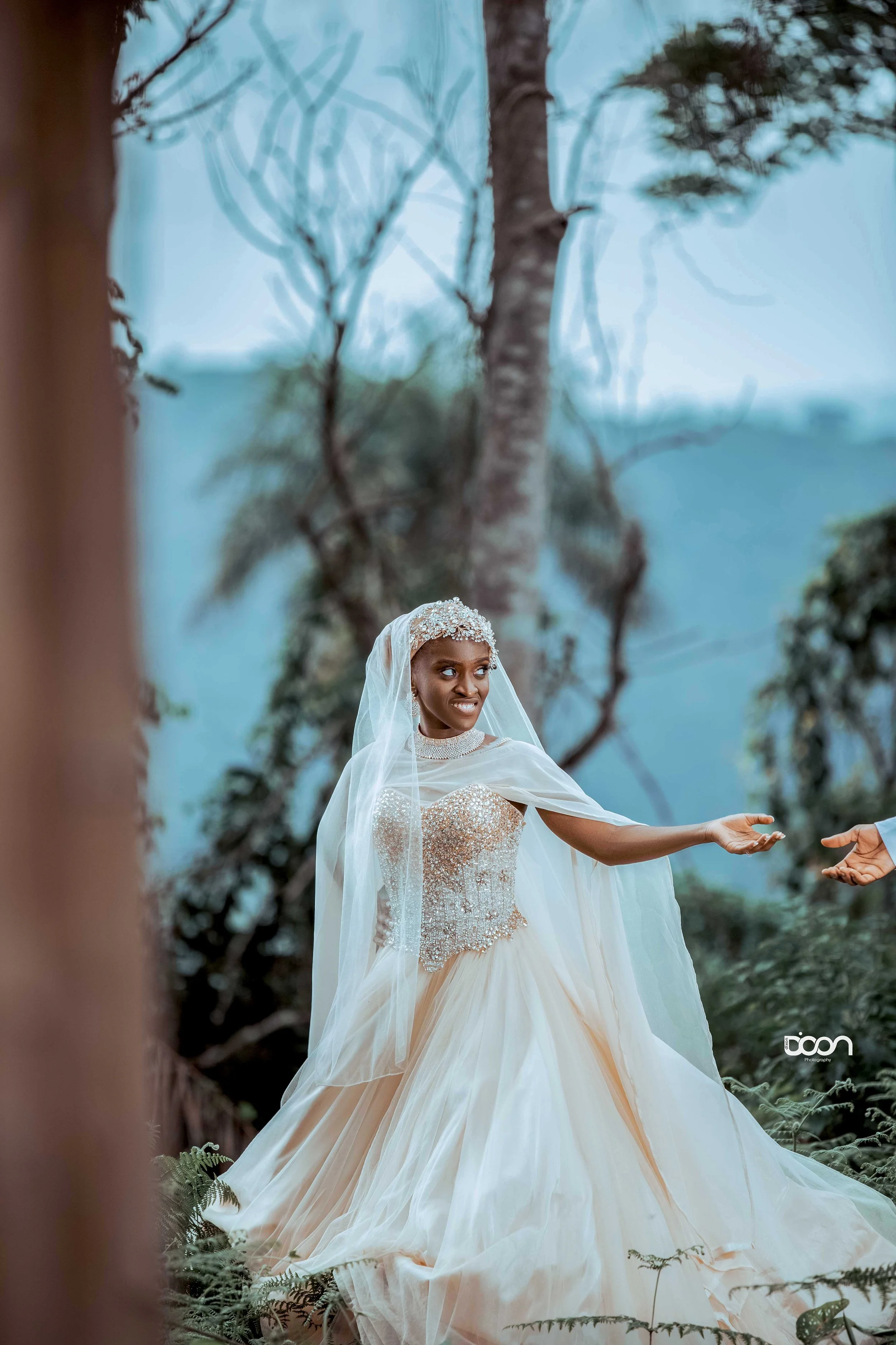 A bride in a strapless beaded wedding gown and veil standing outdoors among trees and greenery, reaching out with her right hand, with a surprised or emotional expression.