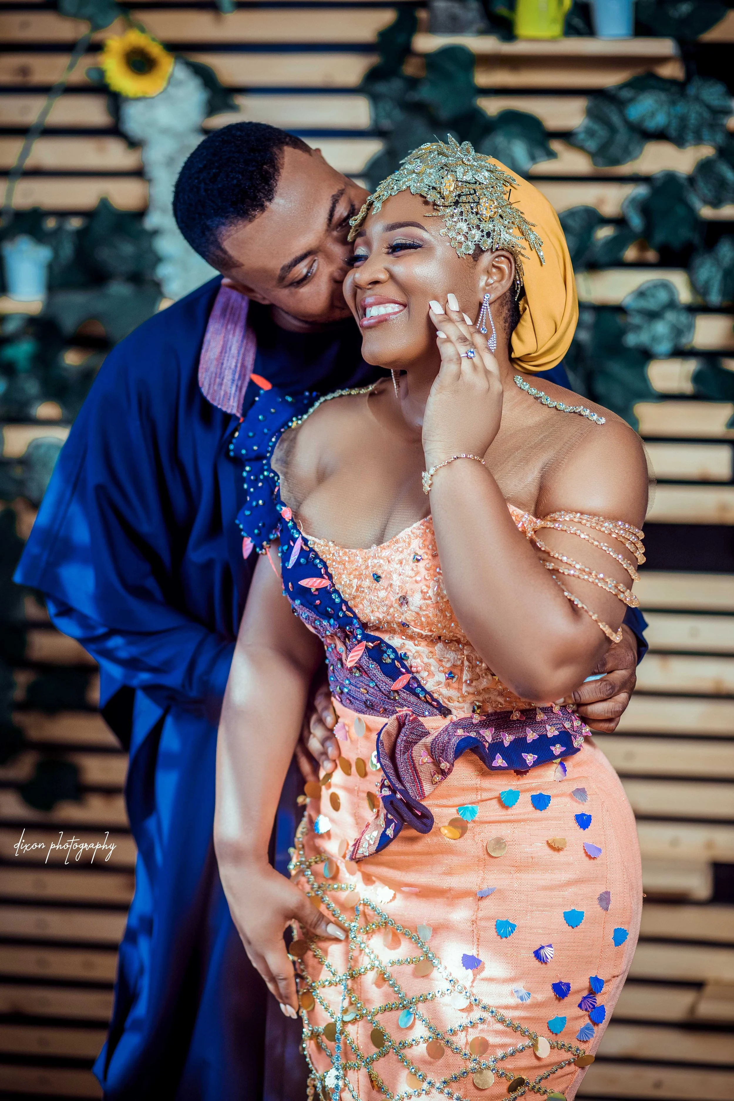 A joyful couple sharing an intimate moment at a celebration, with the woman smiling and touching her face as the man kisses her cheek, both dressed in colorful, embellished attire with a decorative wall and sunflower in the background.