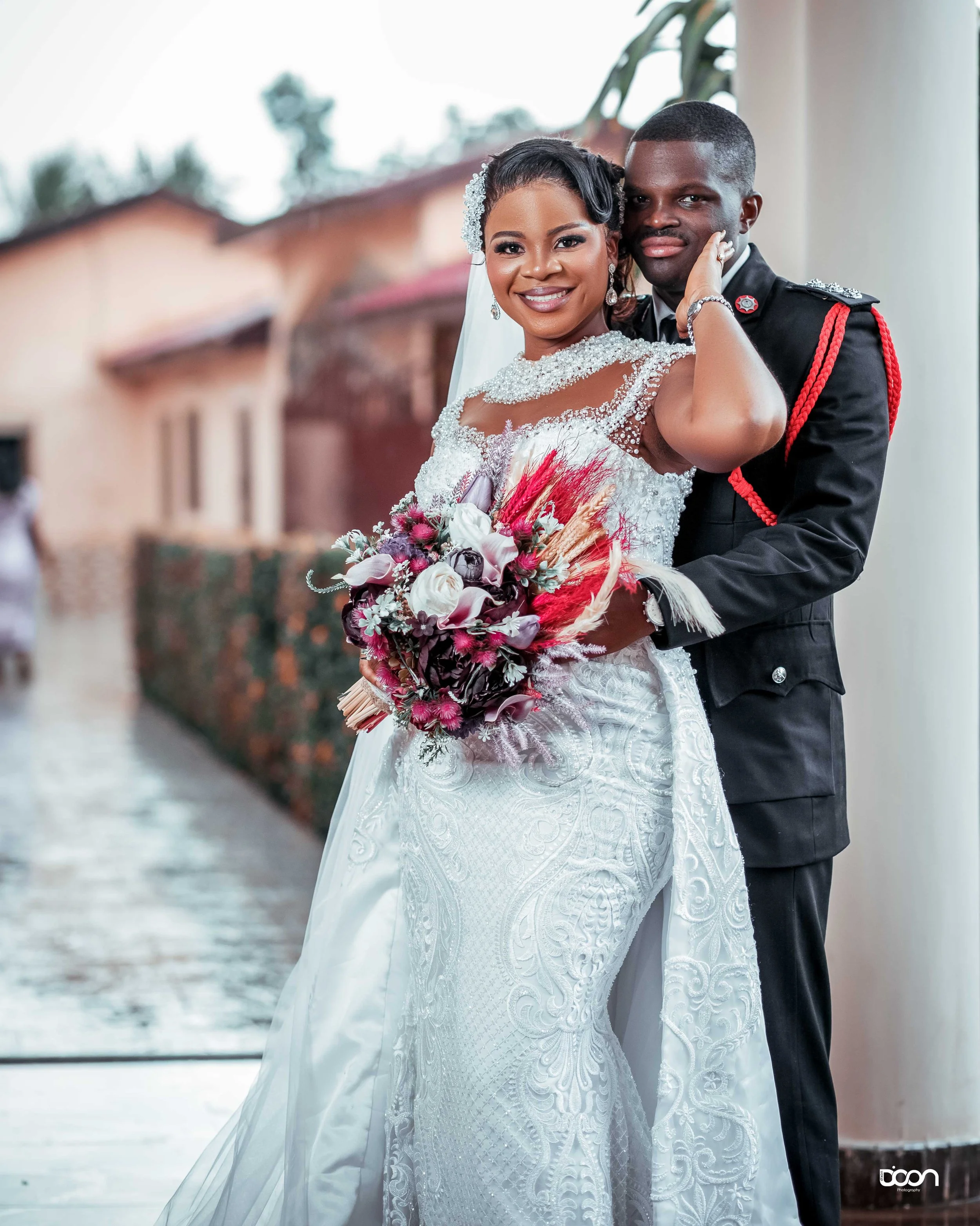 A bride and groom dressed in wedding attire, posing together indoors. The bride is holding a bouquet of flowers and is smiling, while the groom is hugging her from behind.
