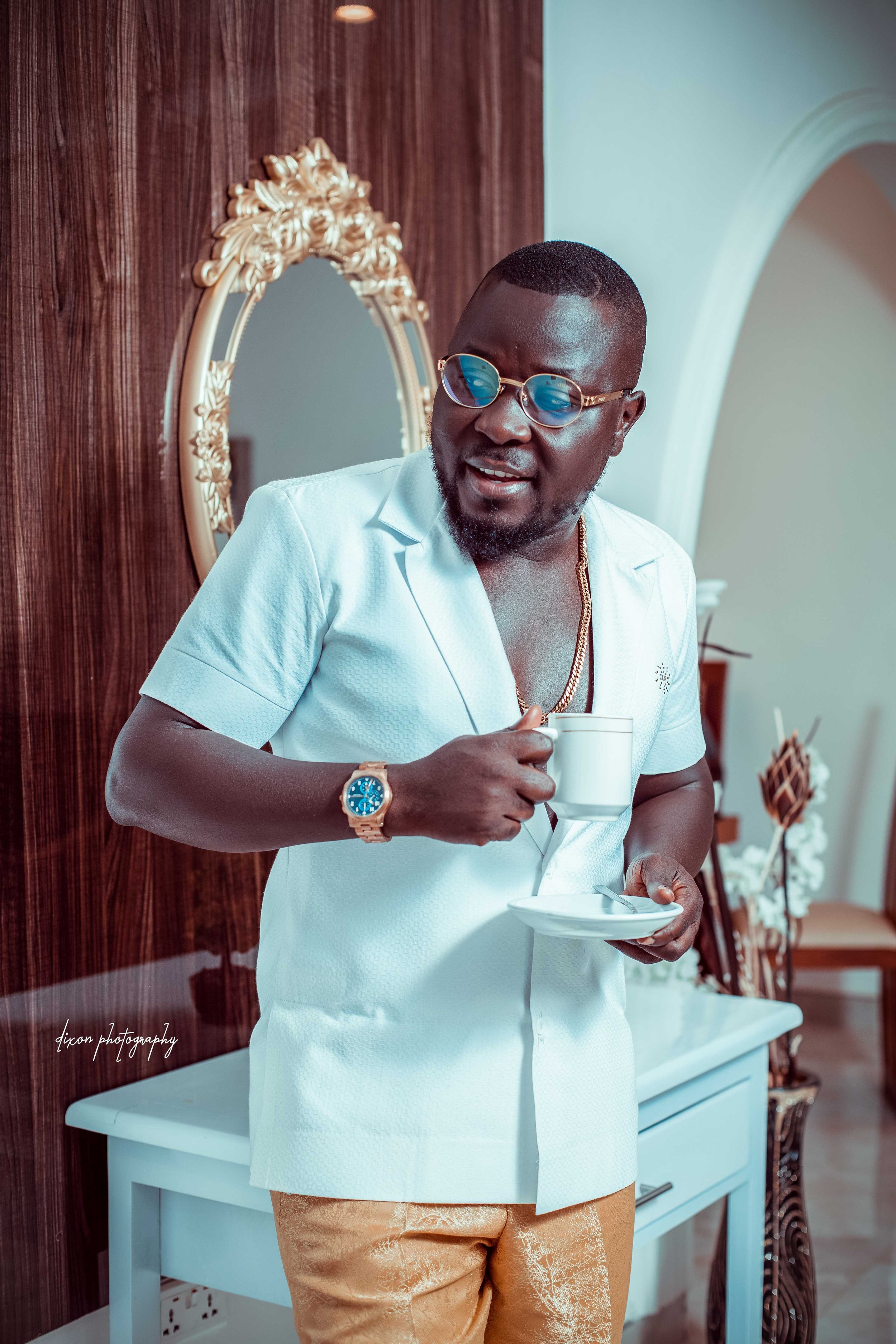 A man wearing sunglasses, a white shirt, gold pants, and a gold watch holding a cup and saucer, standing indoors near a decorative mirror and wooden wall.