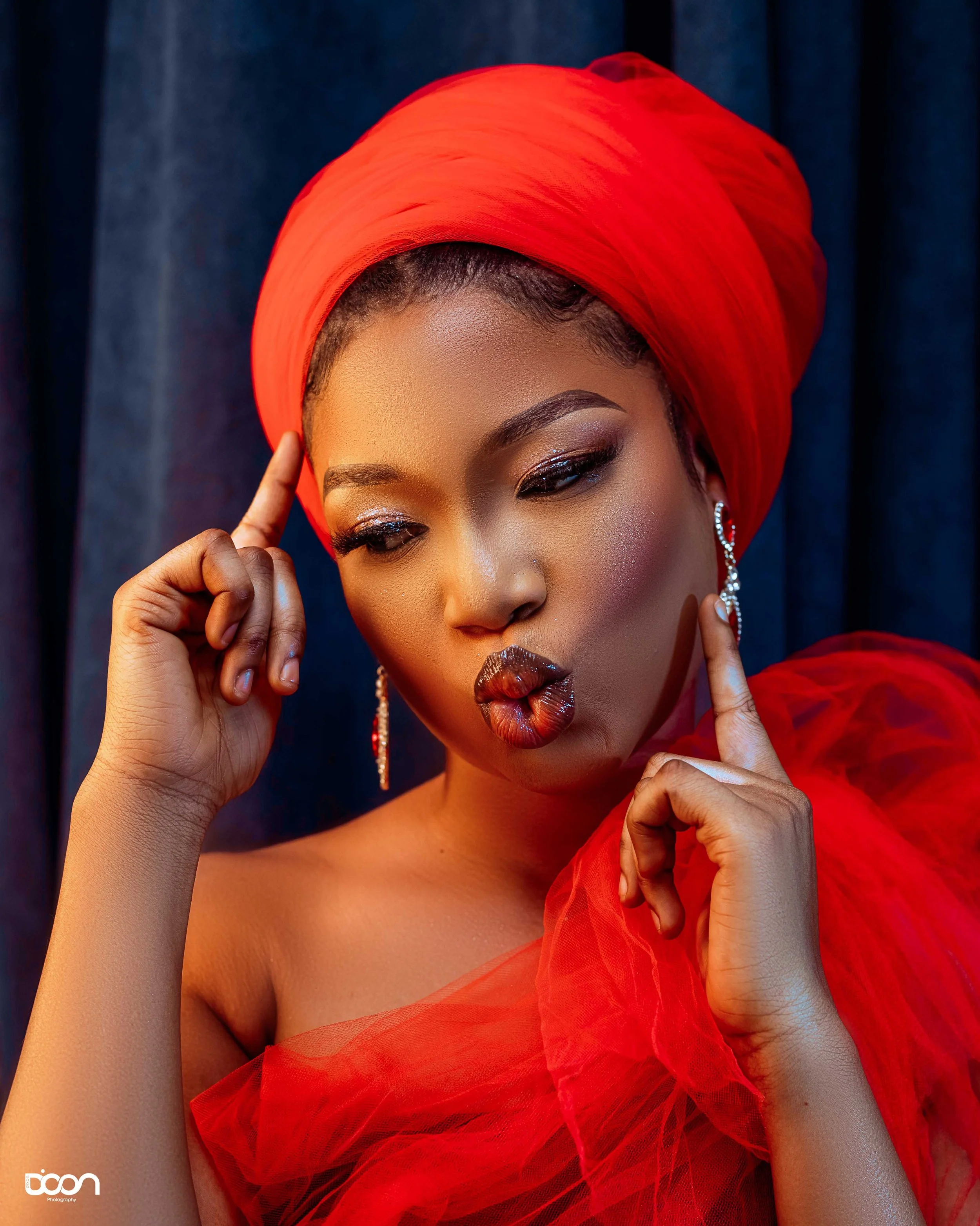 A woman wearing a red headwrap and dress, with makeup and statement earrings, making a kissing face and touching her temples with her fingers.