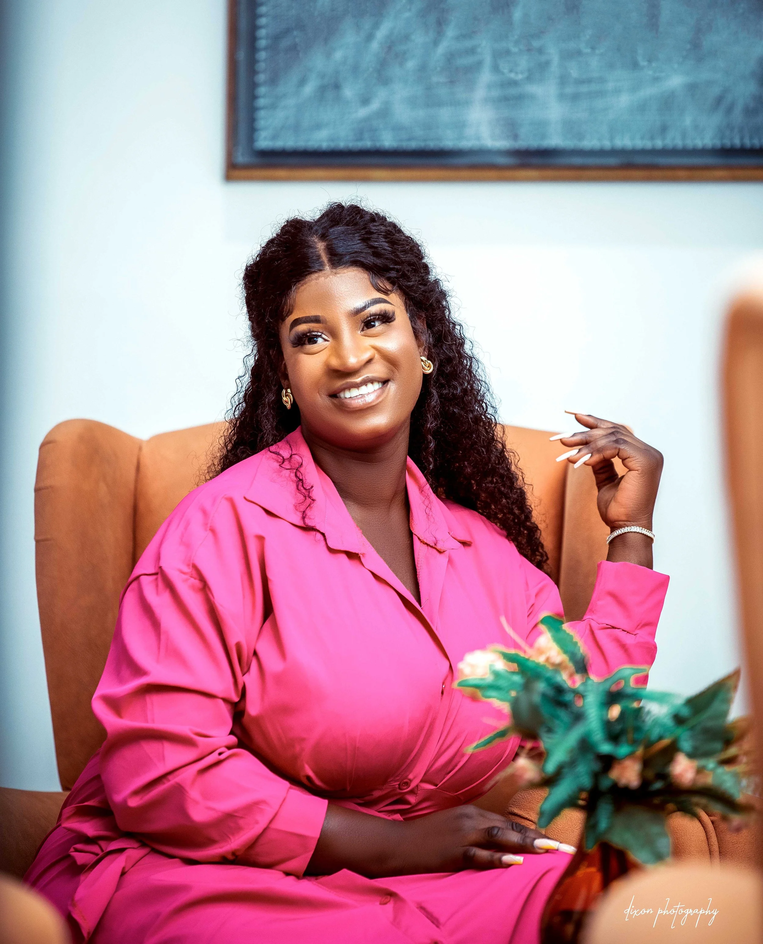 A woman with dark curly hair and a bright smile, wearing a pink dress, sitting on a tan armchair, with a potted plant and a chalkboard in the background.