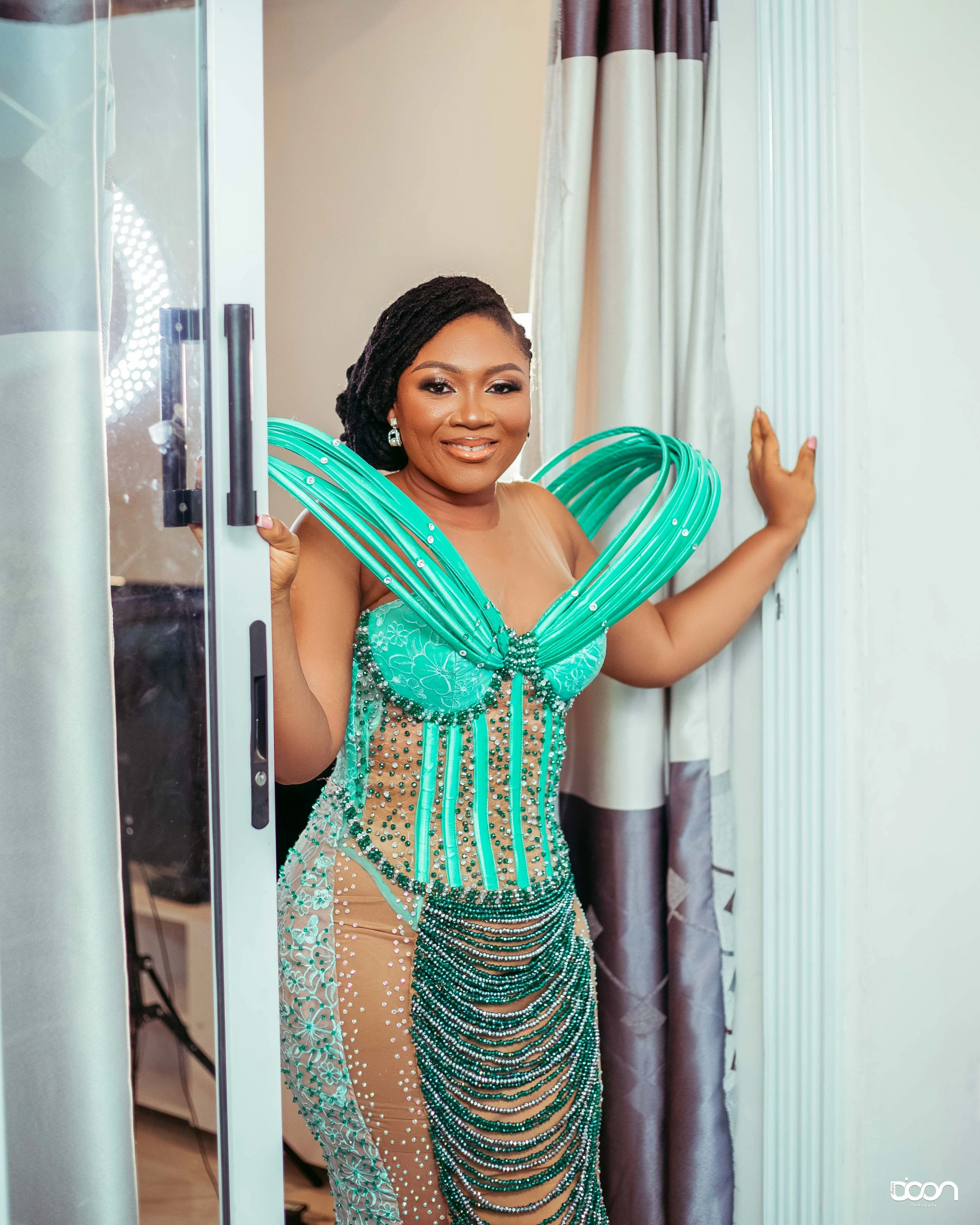 Woman wearing an elaborate turquoise dress with bead and gemstone embellishments, standing in doorway, smiling.