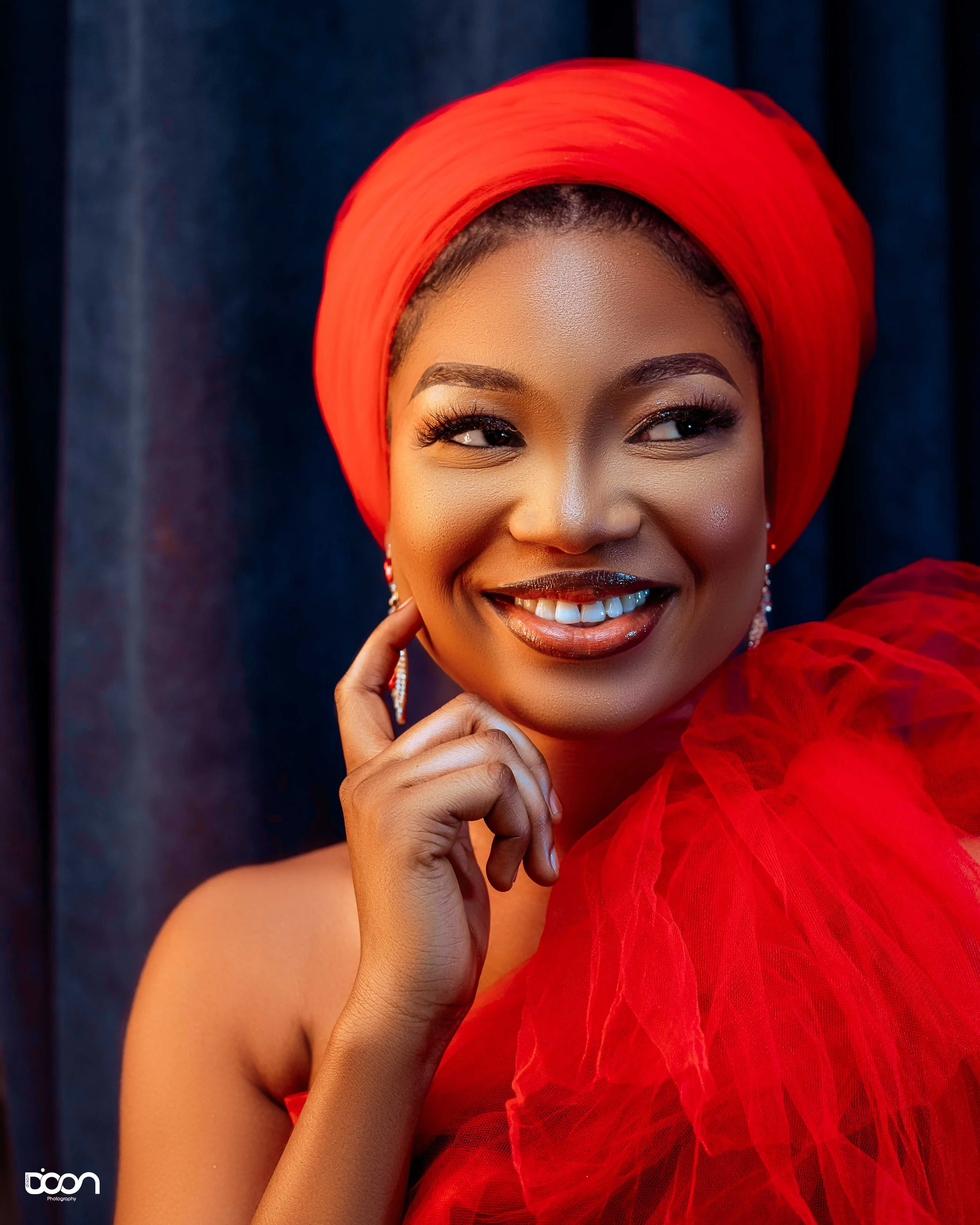 A woman wearing a red headwrap and a red tulle dress smiling with her hand touching her chin, against a dark background.