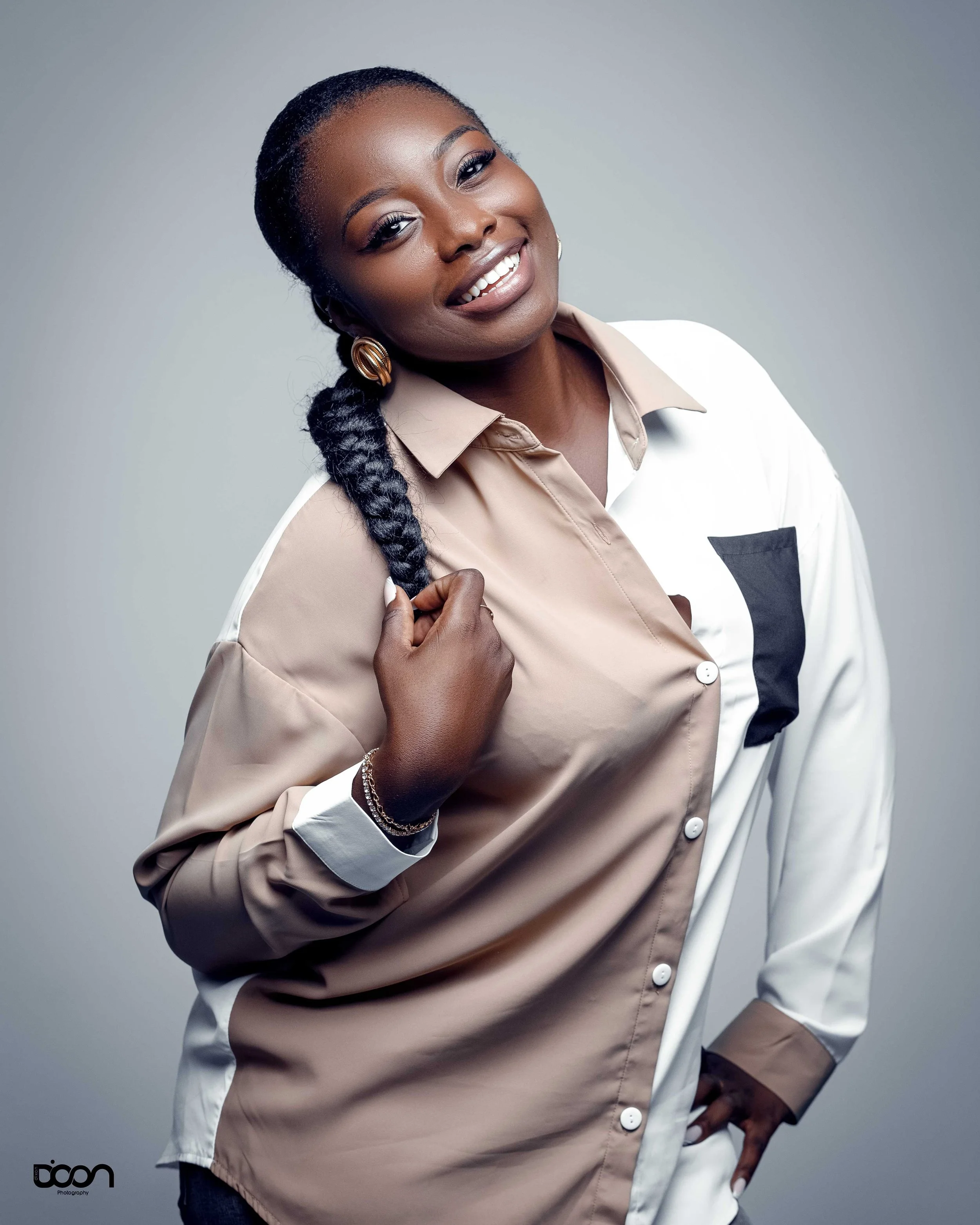 Portrait of a smiling Black woman with long braided hair, wearing a beige and white color-blocked shirt, earrings, bracelet, and holding her braid with one hand against a plain grey background.