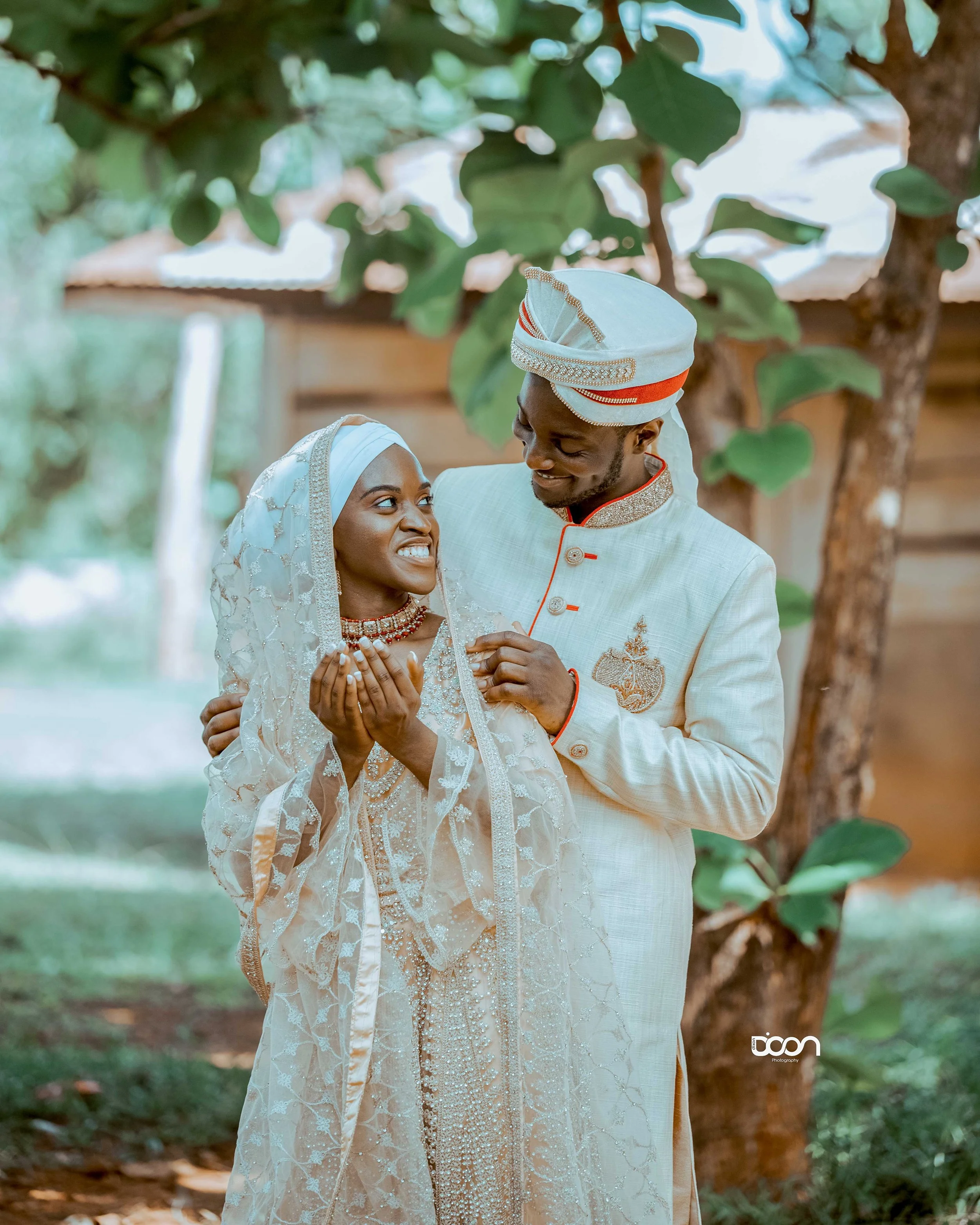 A couple in traditional African wedding attire standing under a tree, smiling and gazing at each other.