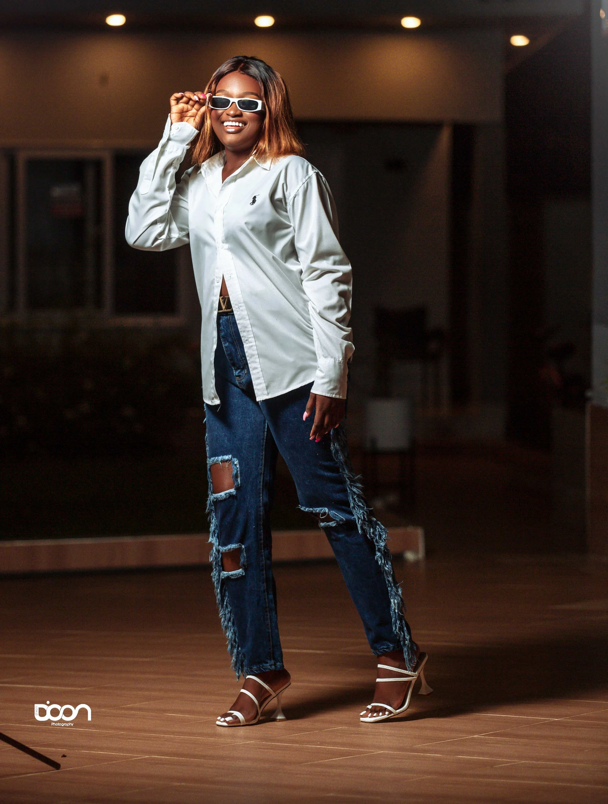 Woman wearing sunglasses, oversized shirt, ripped jeans with fringe, and high heels, standing indoors at night, smiling and adjusting her glasses.