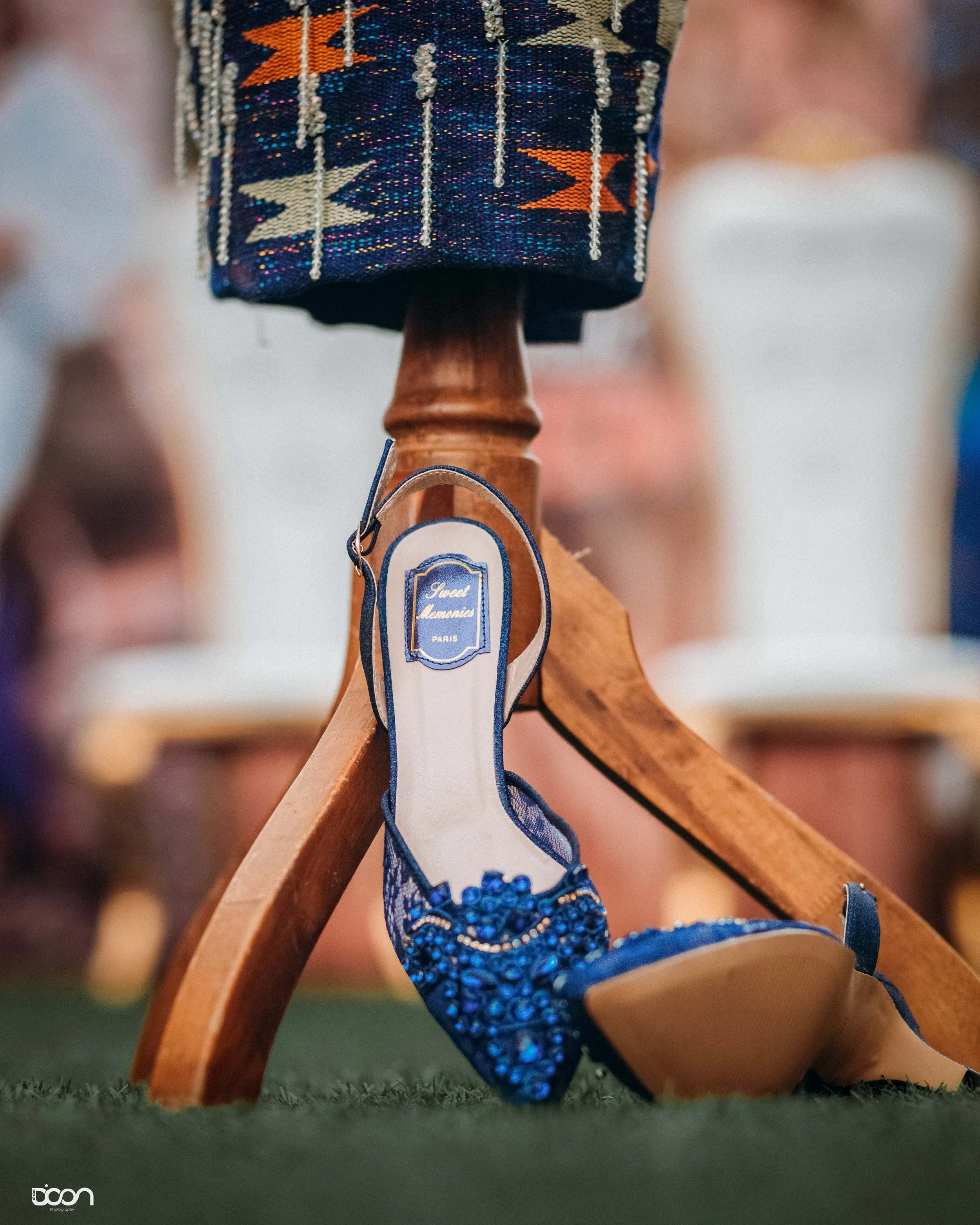 A blue jeweled high-heeled shoe hanging from a wooden furniture leg, with a fabric with colorful beaded patterns draped over the furniture. The shoe has a tag that reads "Sweet Memories PARIS".