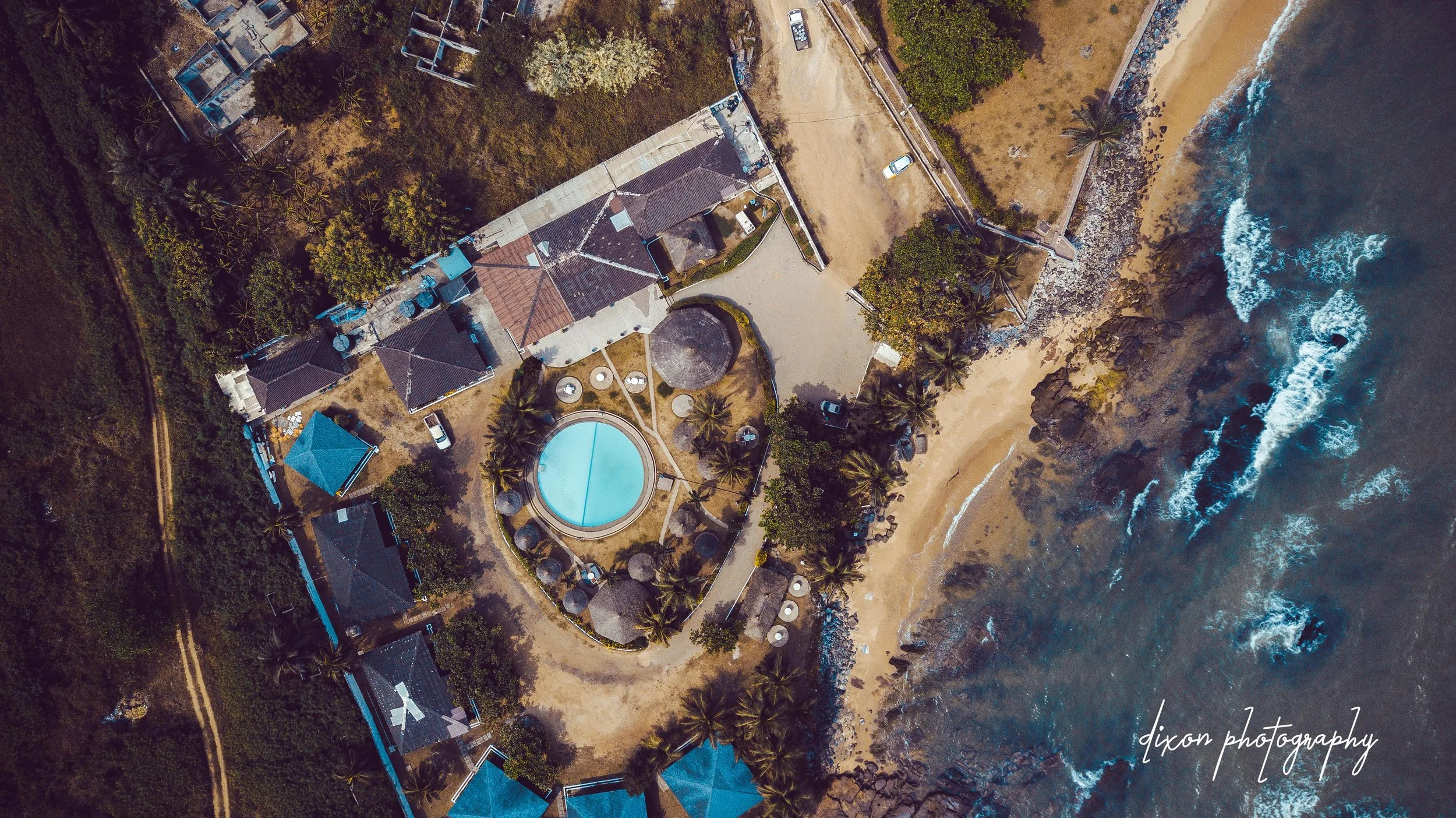 Aerial view of a beachfront resort with a circular swimming pool, surrounded by palm trees, thatched-roof structures, and a sandy beach with waves on the shoreline.