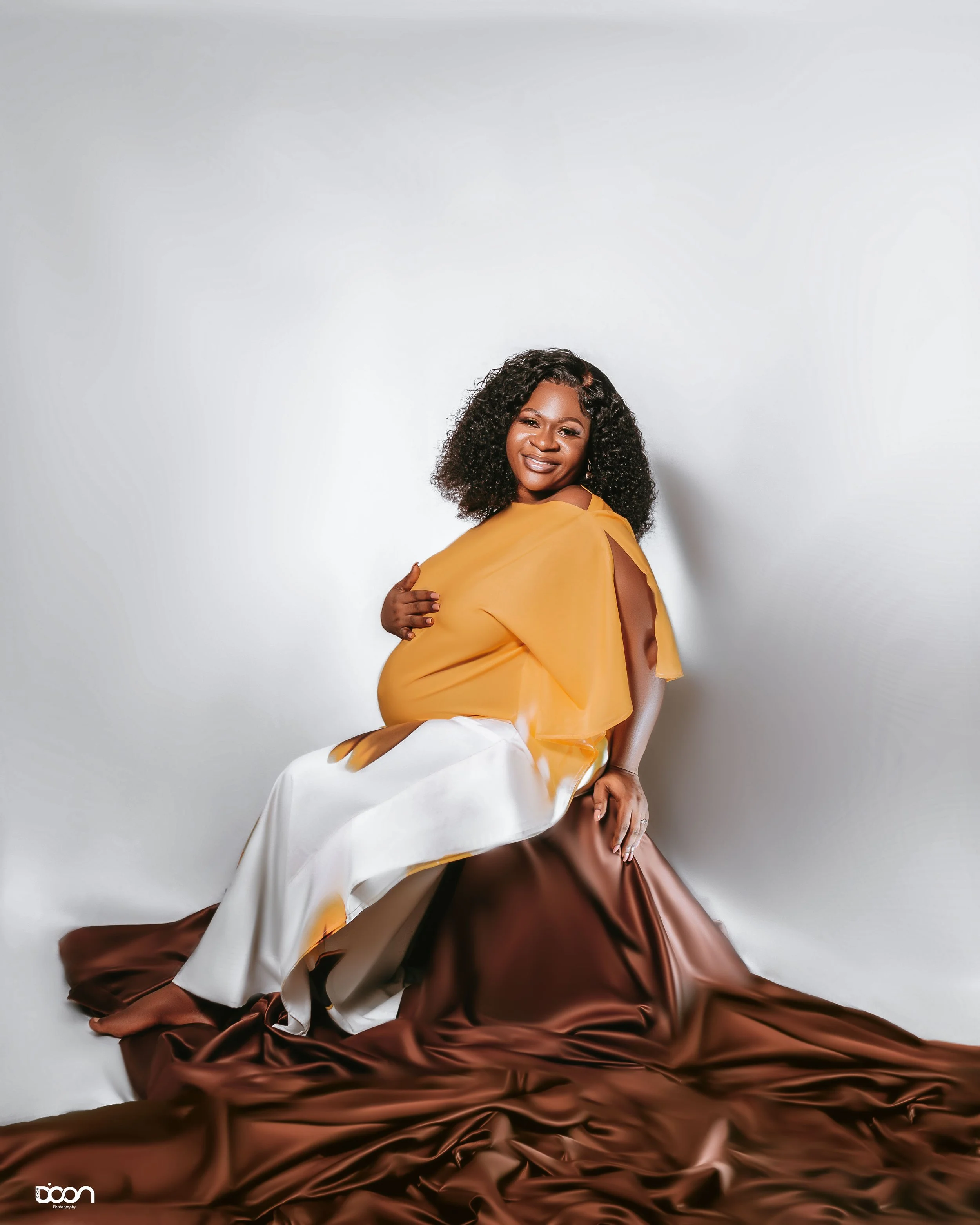 A pregnant woman with curly hair sitting on a satin brown fabric, wearing a yellow top and white pants, smiling at the camera against a plain white background.