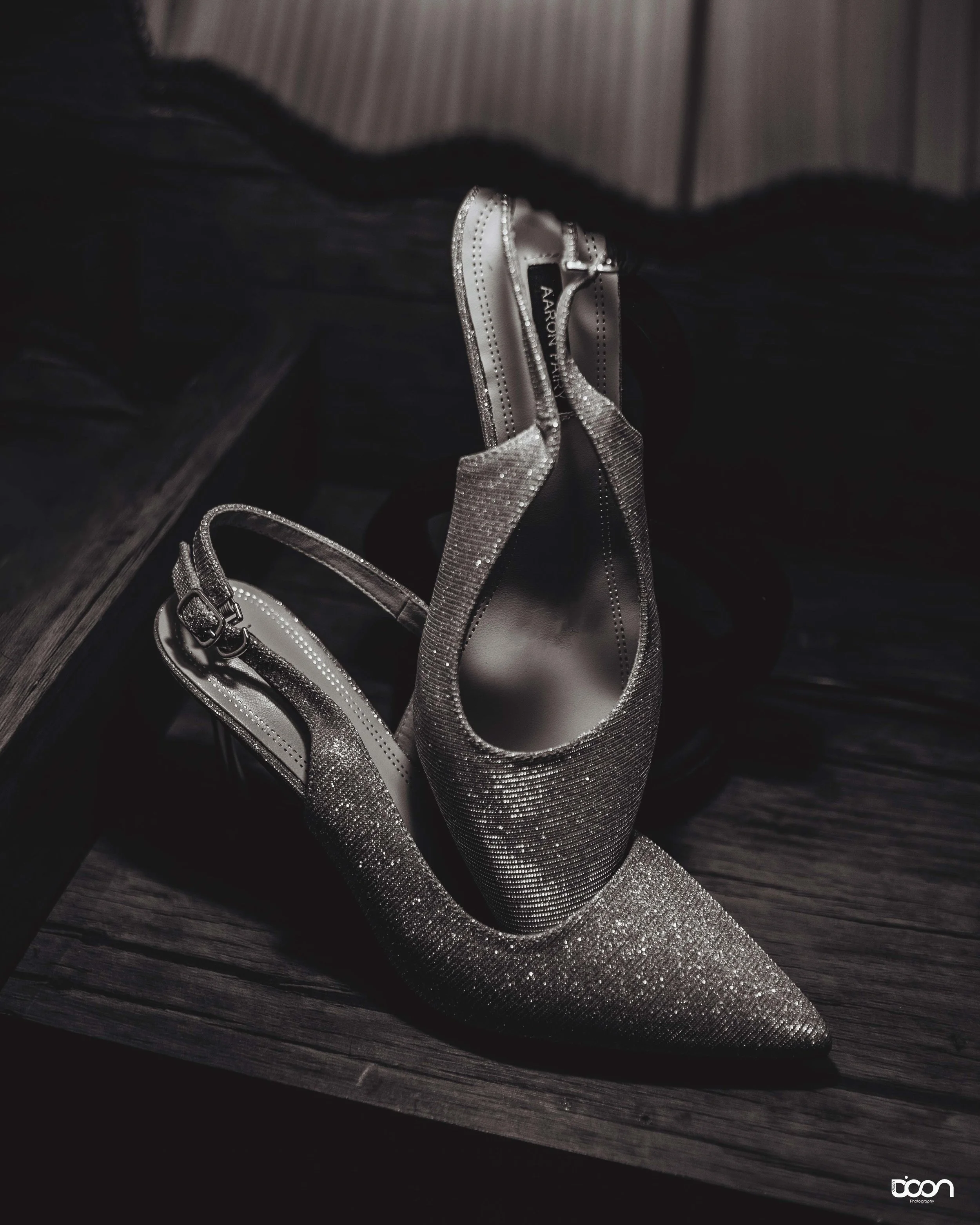 A pair of sparkling, silver high-heeled shoes with pointed toes and ankle straps, resting on a dark wooden surface.