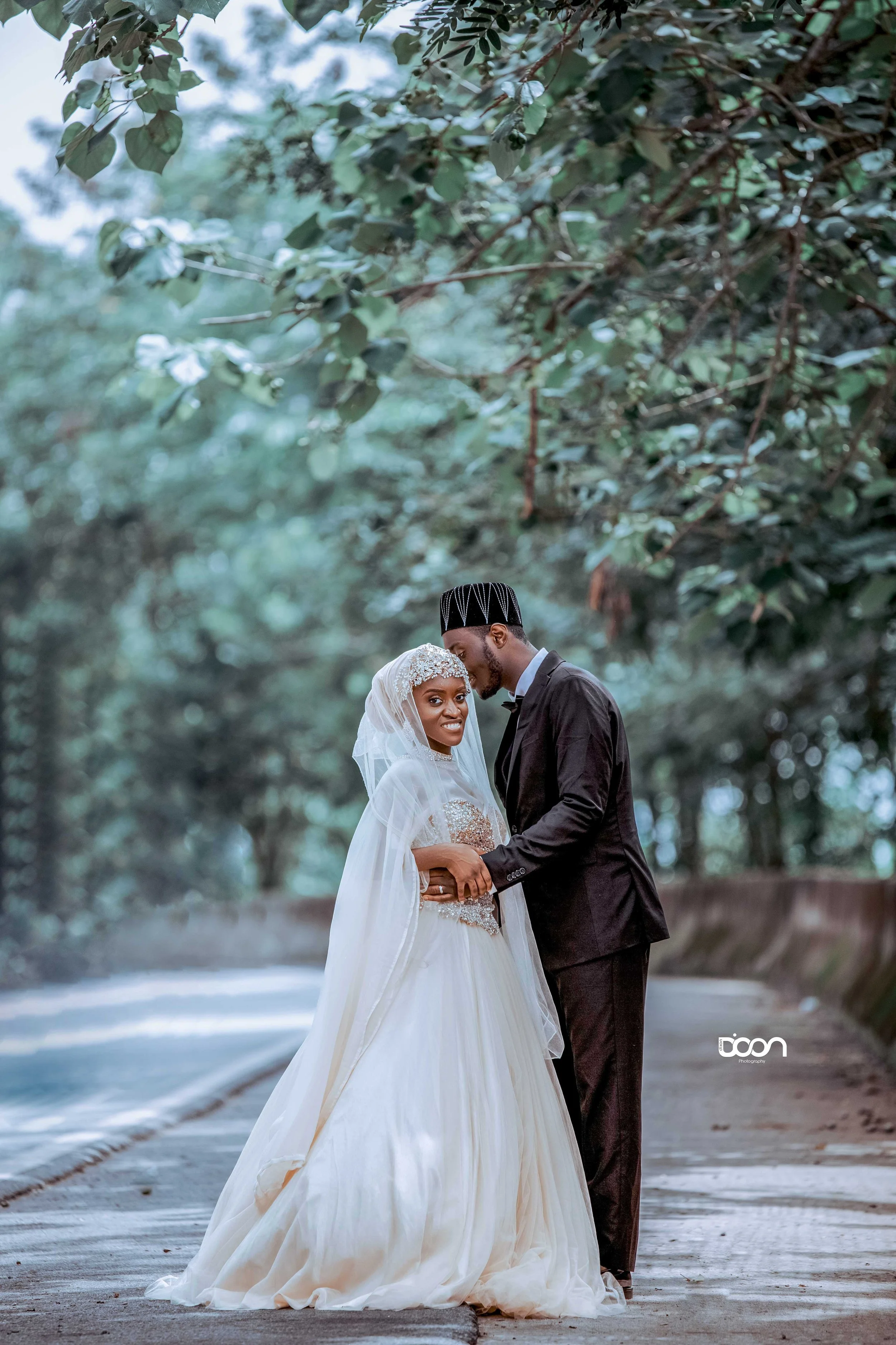 A wedding couple standing on a pathway surrounded by trees, with the groom leaning his forehead against the bride's. The bride is wearing a white gown with intricate detailing, a veil, and a headpiece, while the groom is dressed in a dark suit and tr