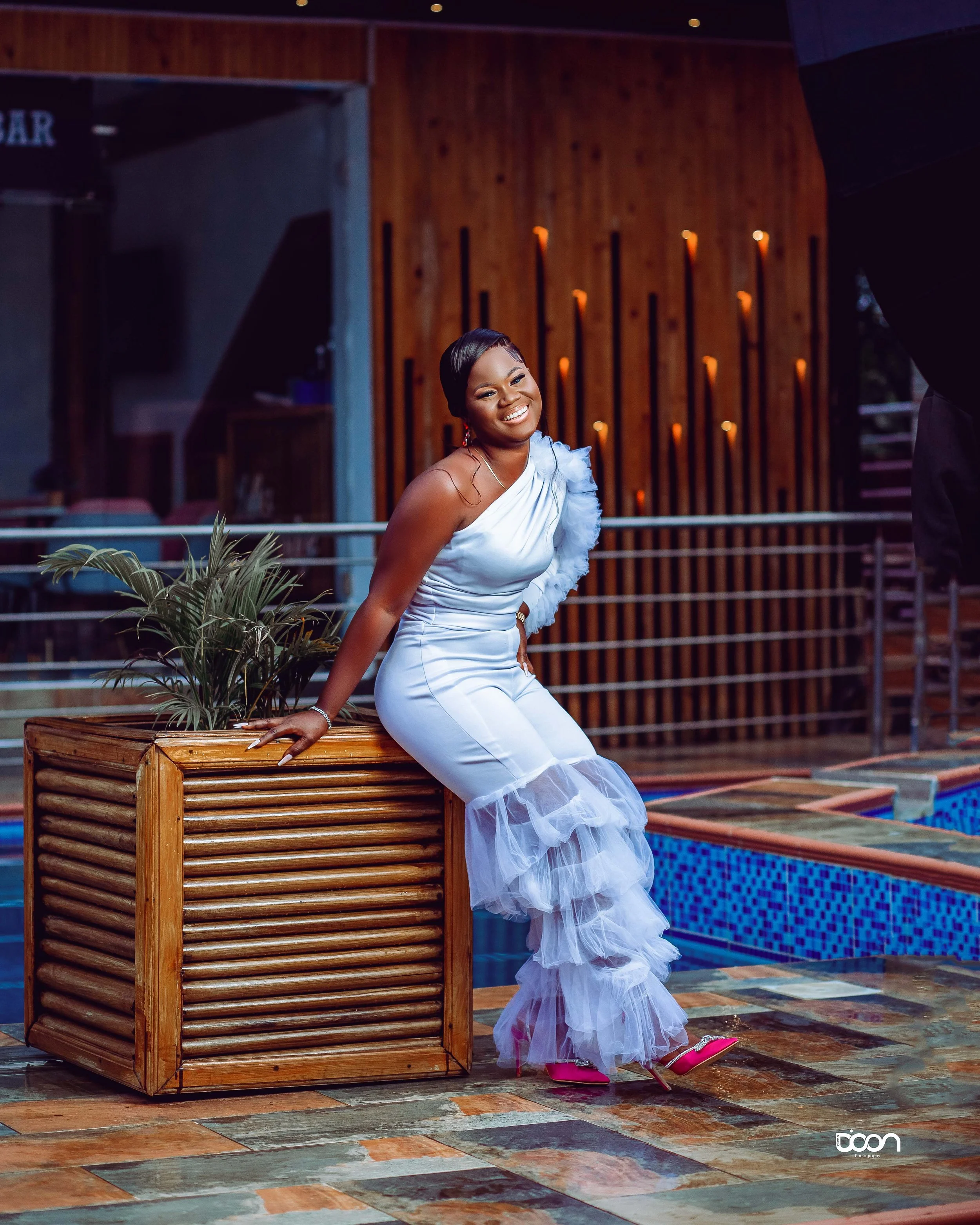 A woman in a white dress with ruffled sheer accents, pink high heels, and earrings sitting on a wooden planter box with a plant, smiling and posing at a colorful poolside area with wooden wall background.