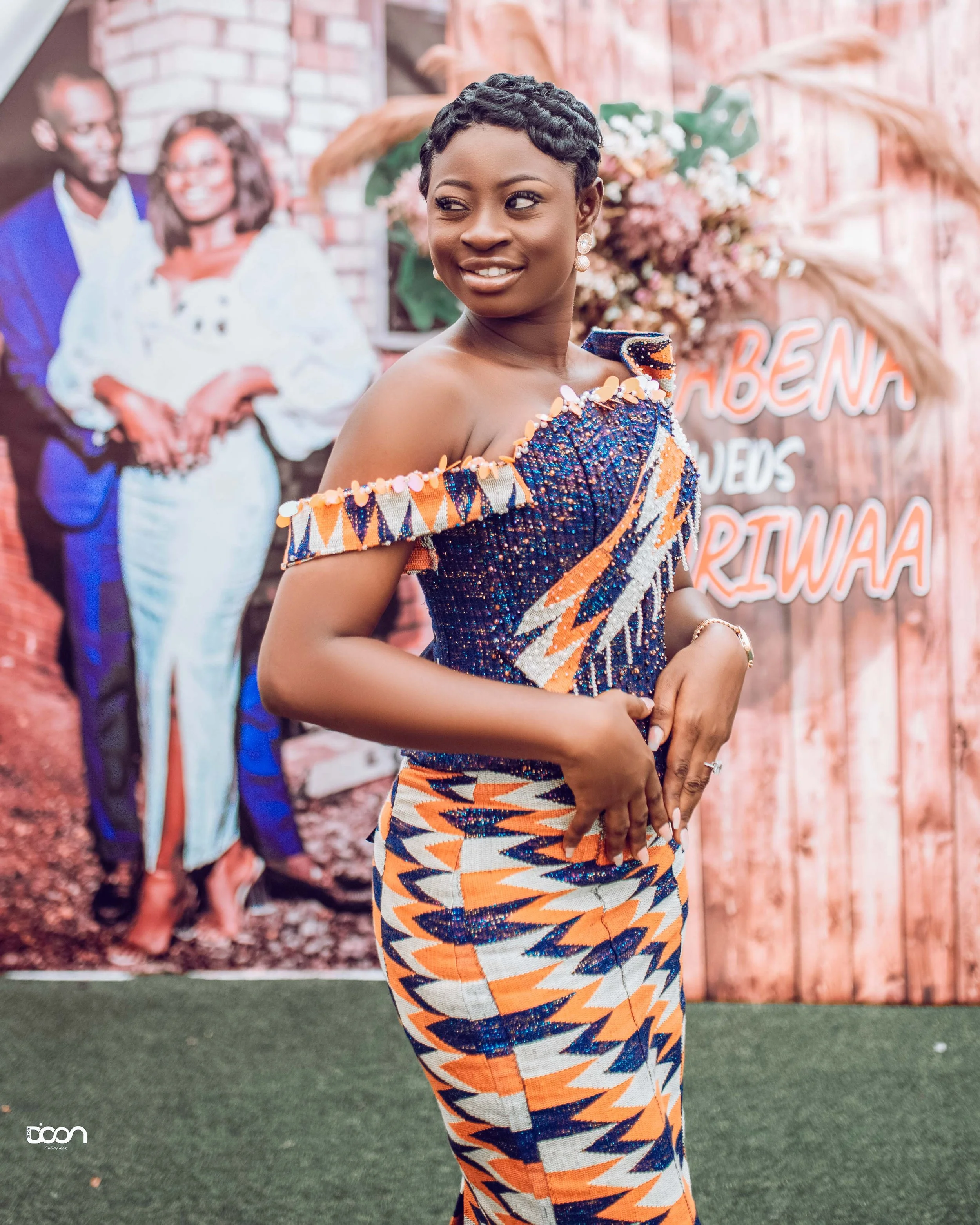 A woman dressed in traditional African attire, standing indoors with a smile, in front of a backdrop that features a large photo of a couple and a wooden wall with floral decorations. The background includes cursive lettering that reads 'NABENA' and 
