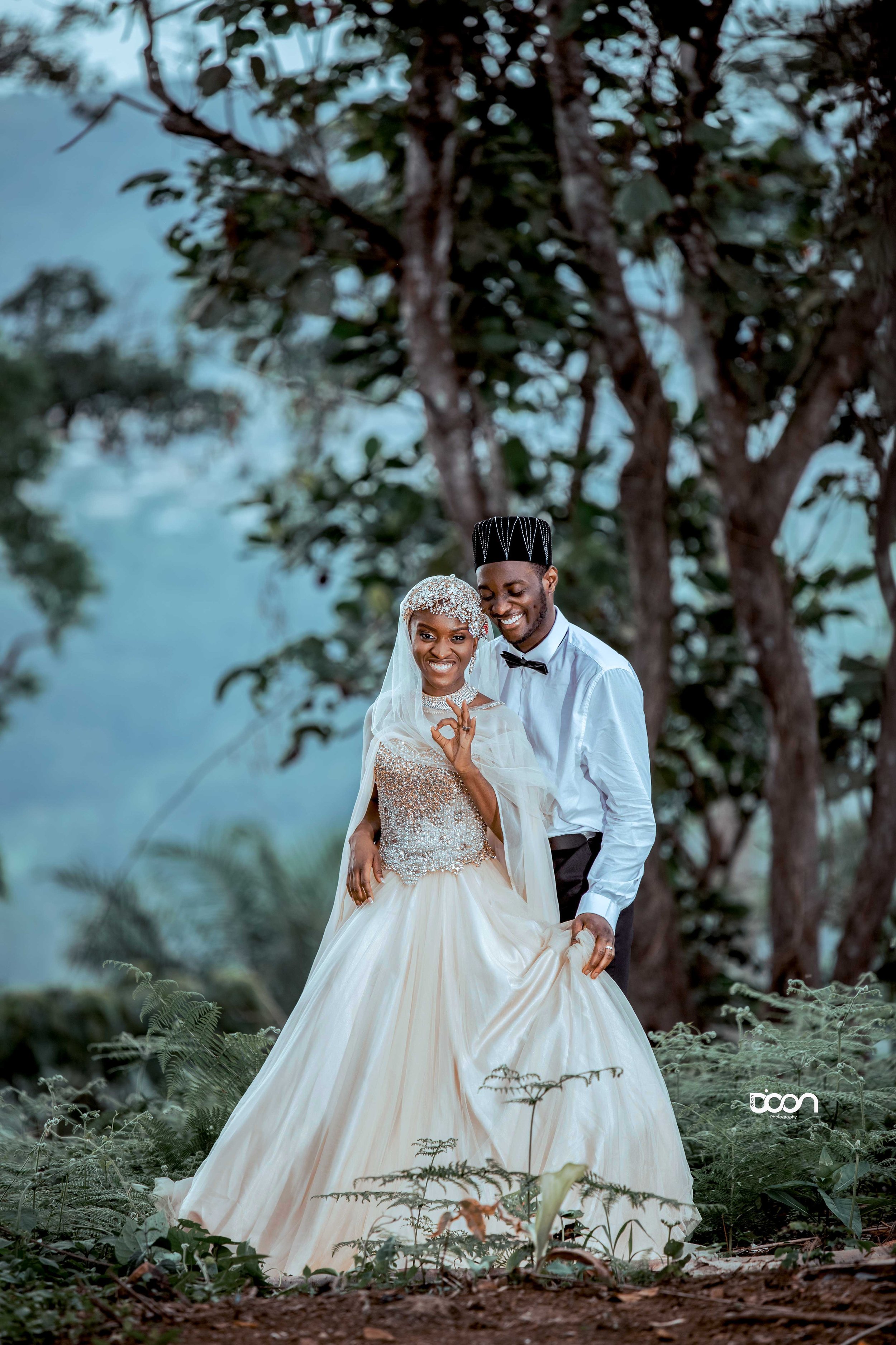 A happy couple dressed in wedding attire, standing outdoors among trees and plants, smiling and making a heart shape with their hands.