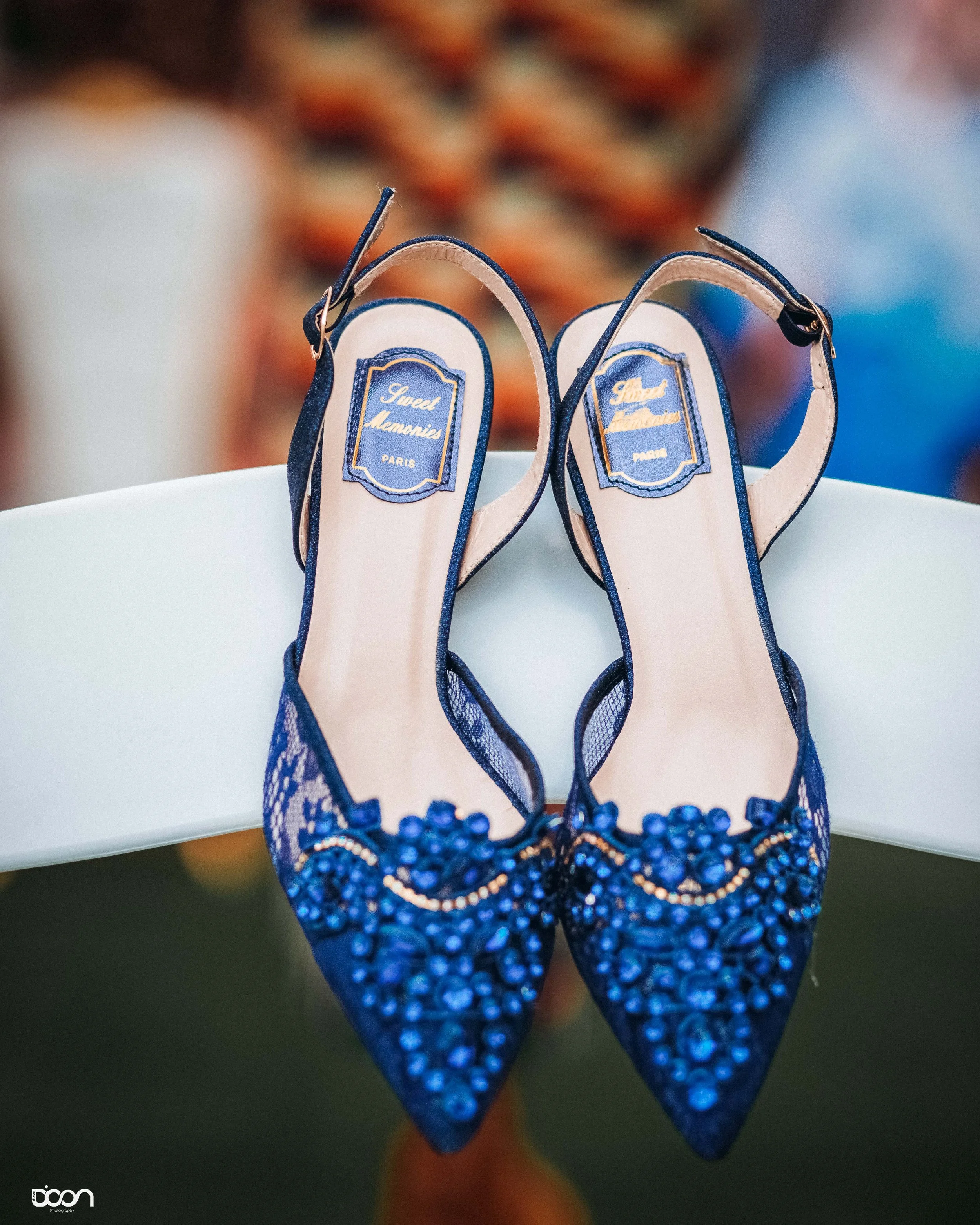 A pair of elegant blue high-heeled shoes with embellishments rests on a white surface, with a blurred patterned background.