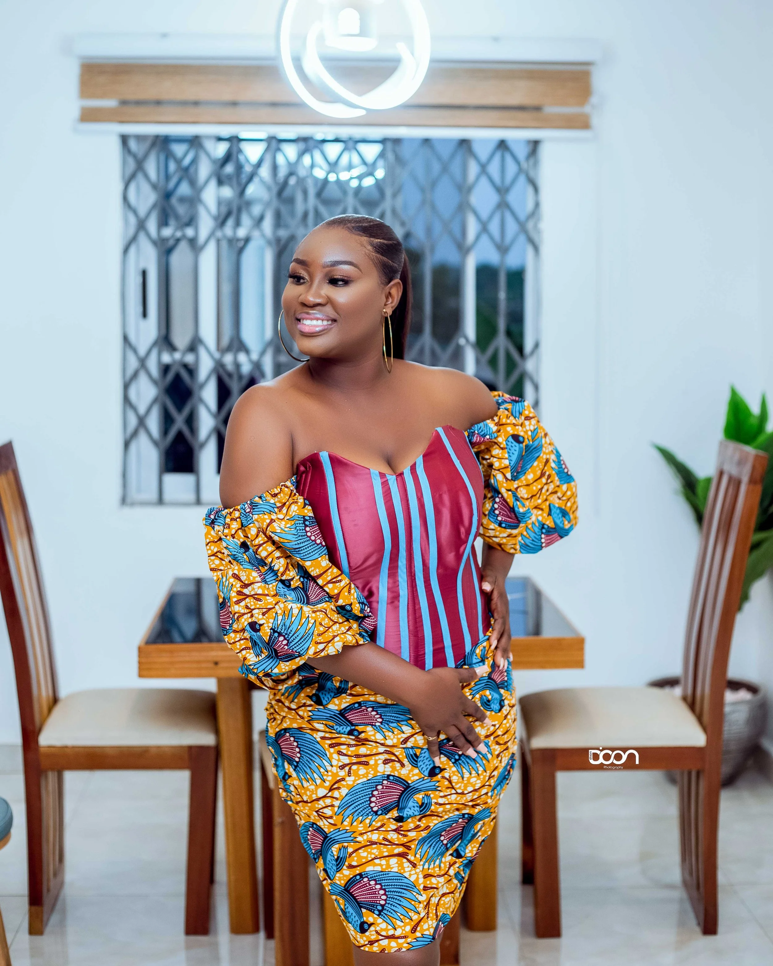 A woman with dark skin smiling and looking to the side, wearing a colorful off-shoulder dress with blue and yellow African print patterns and a corset-style top with red and pink vertical stripes, standing in a room with wooden chairs and green plant