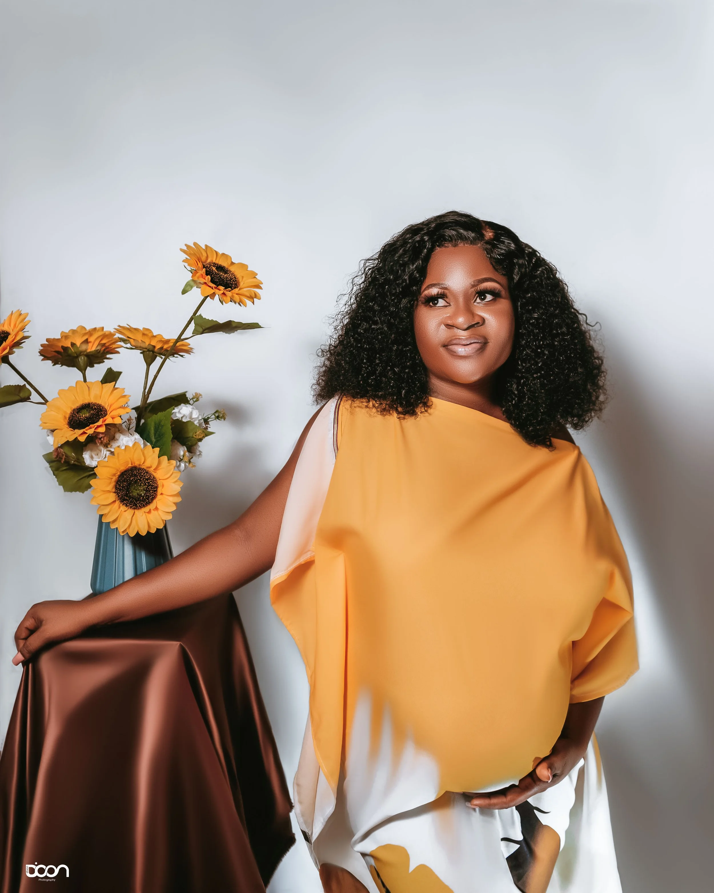 A woman with curly hair wearing a yellow top and white pants, standing next to a table with a vase of sunflowers, against a plain light-colored background.