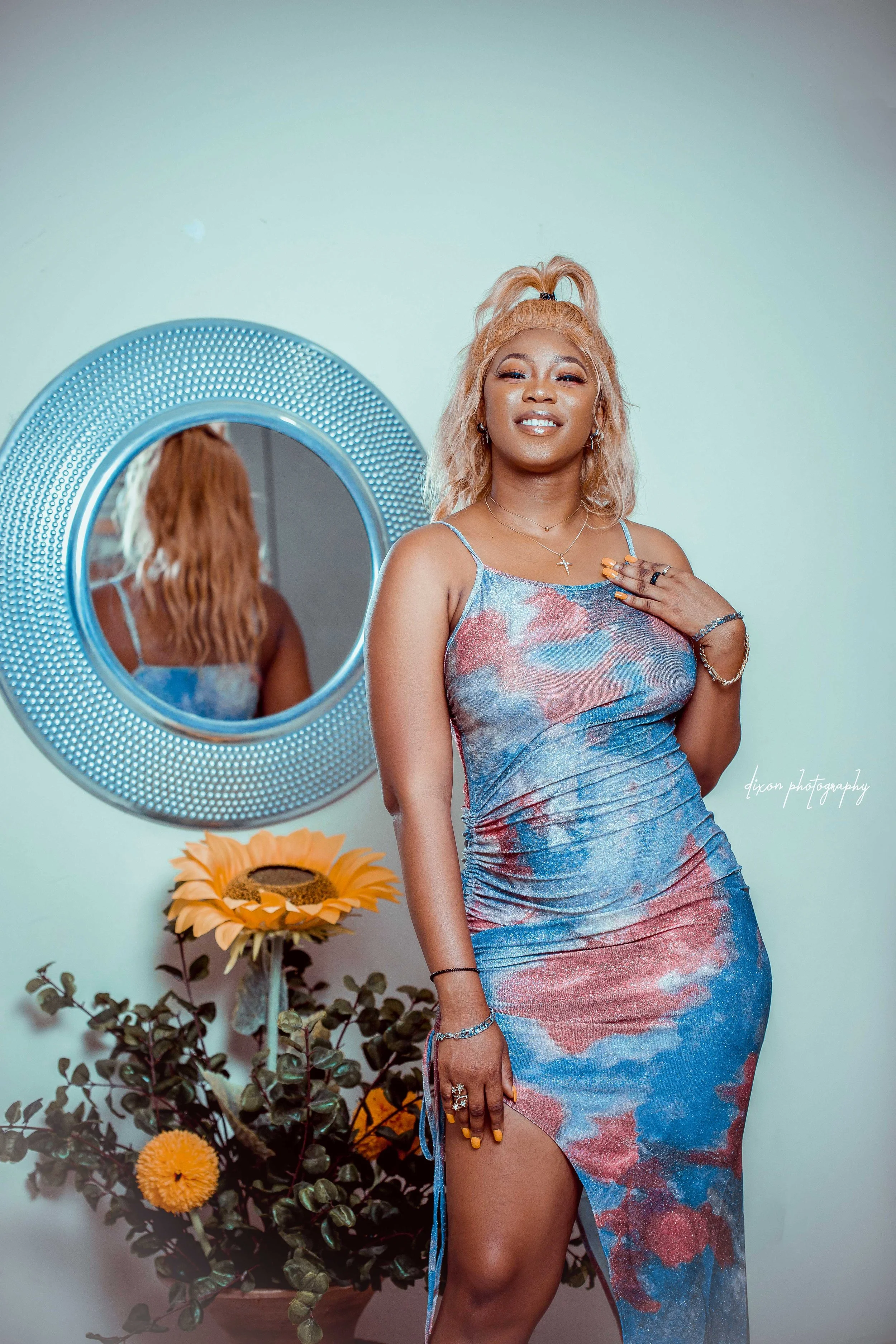 A young woman with blonde curly hair wearing a colorful tie-dye dress standing near a round mirror, with a sunflower plant and yellow-orange flowers below, smiling with one hand on her chest.