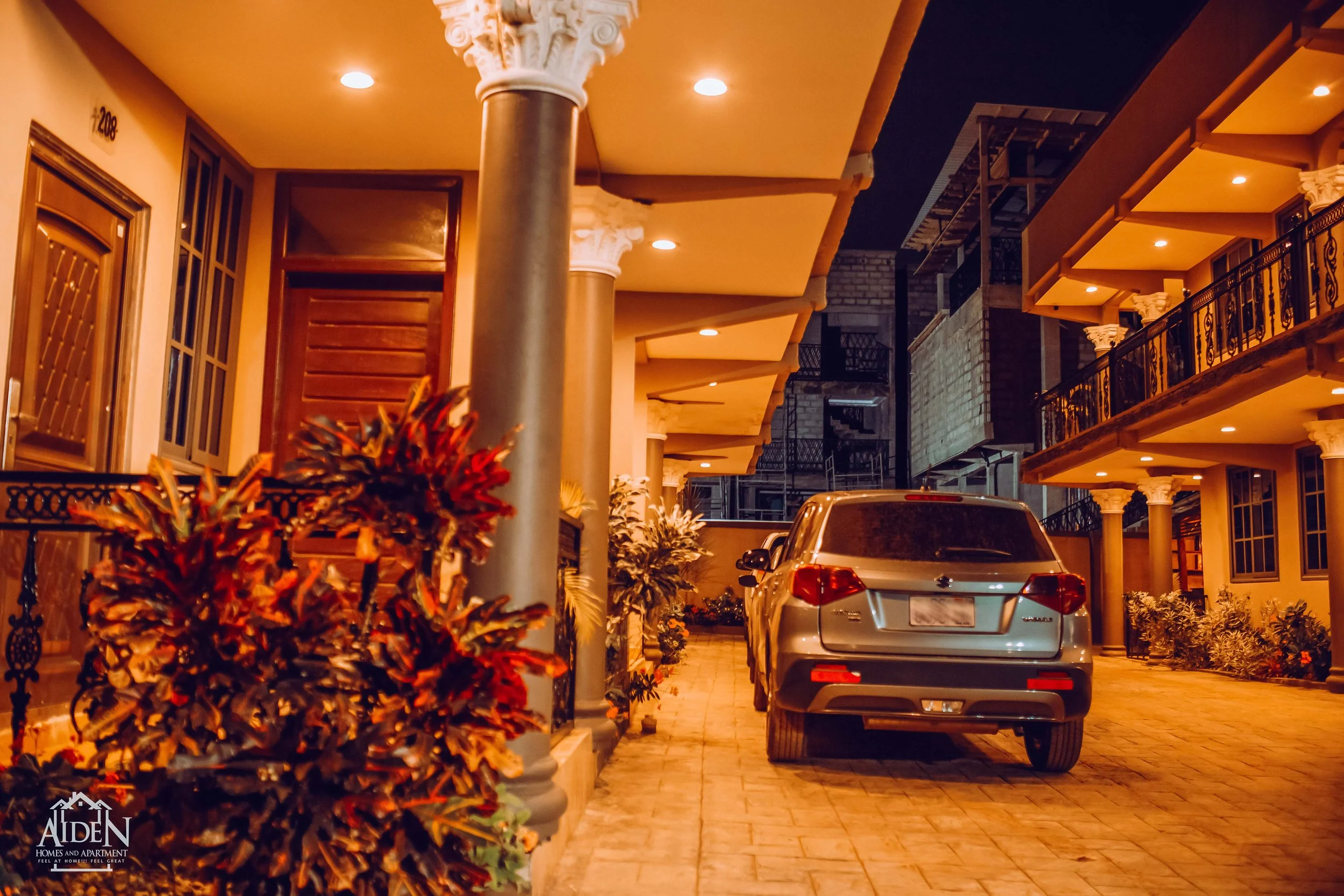 Nighttime view of a residential building with a parking area. There is a silver SUV parked, illuminated by warm exterior lighting. The building has arched columns, balconies with decorative railings, and lush plants along the pathway.