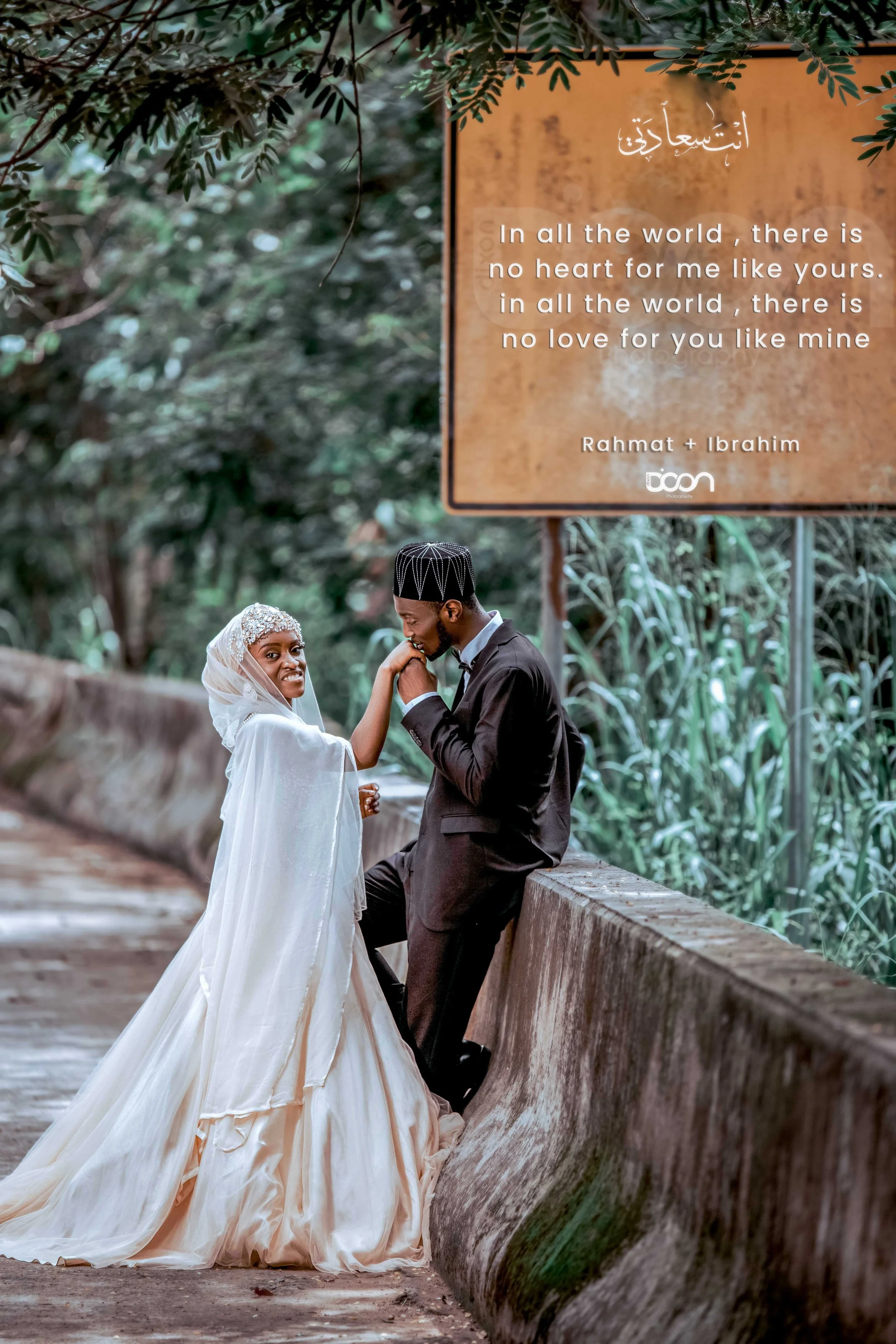 A bride and groom in wedding attire outdoors, with the bride touching the groom's chin, and a large sign with romantic quote in the background.