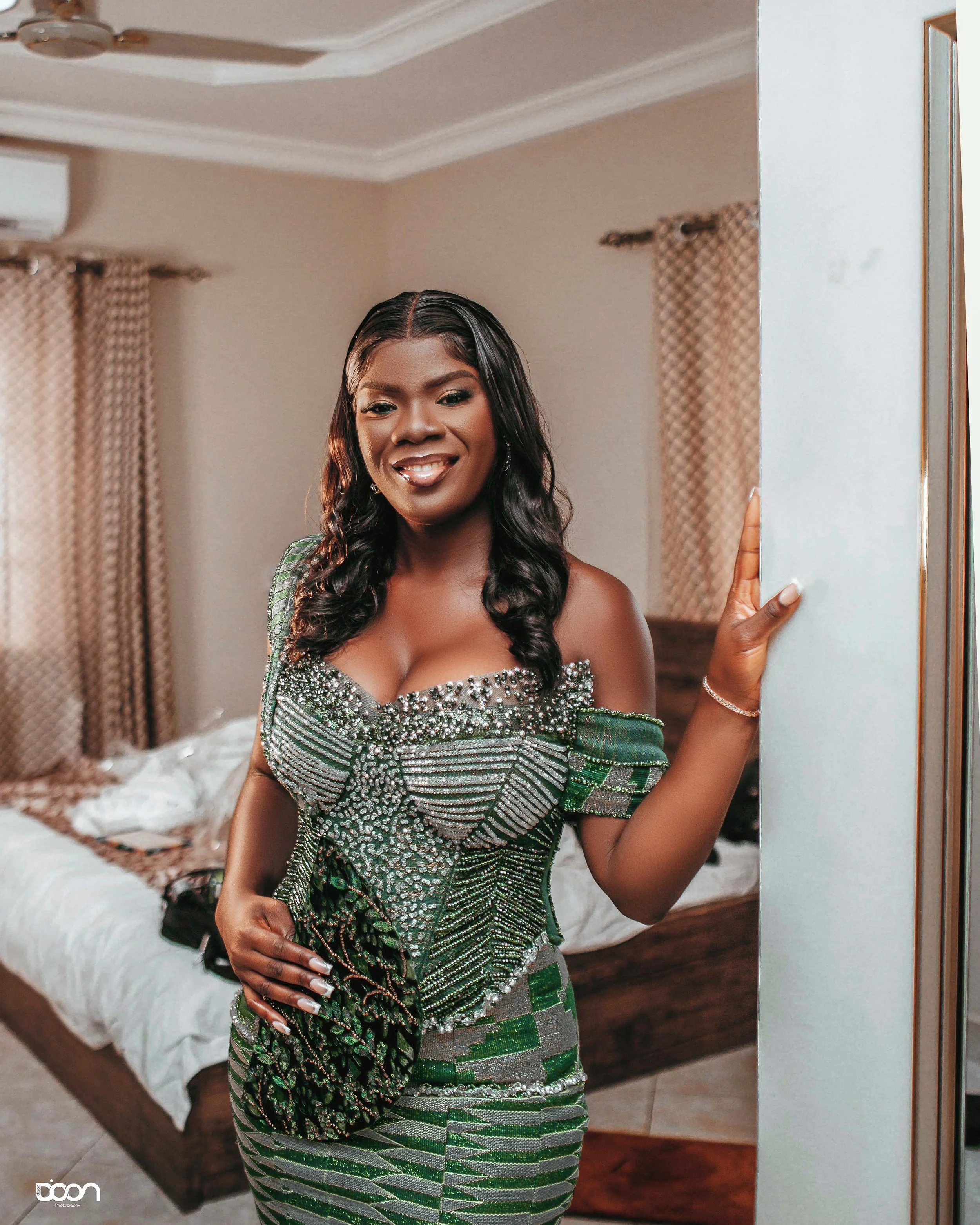 A woman dressed in a green and silver beaded dress, smiling and standing by a wall in a bedroom.