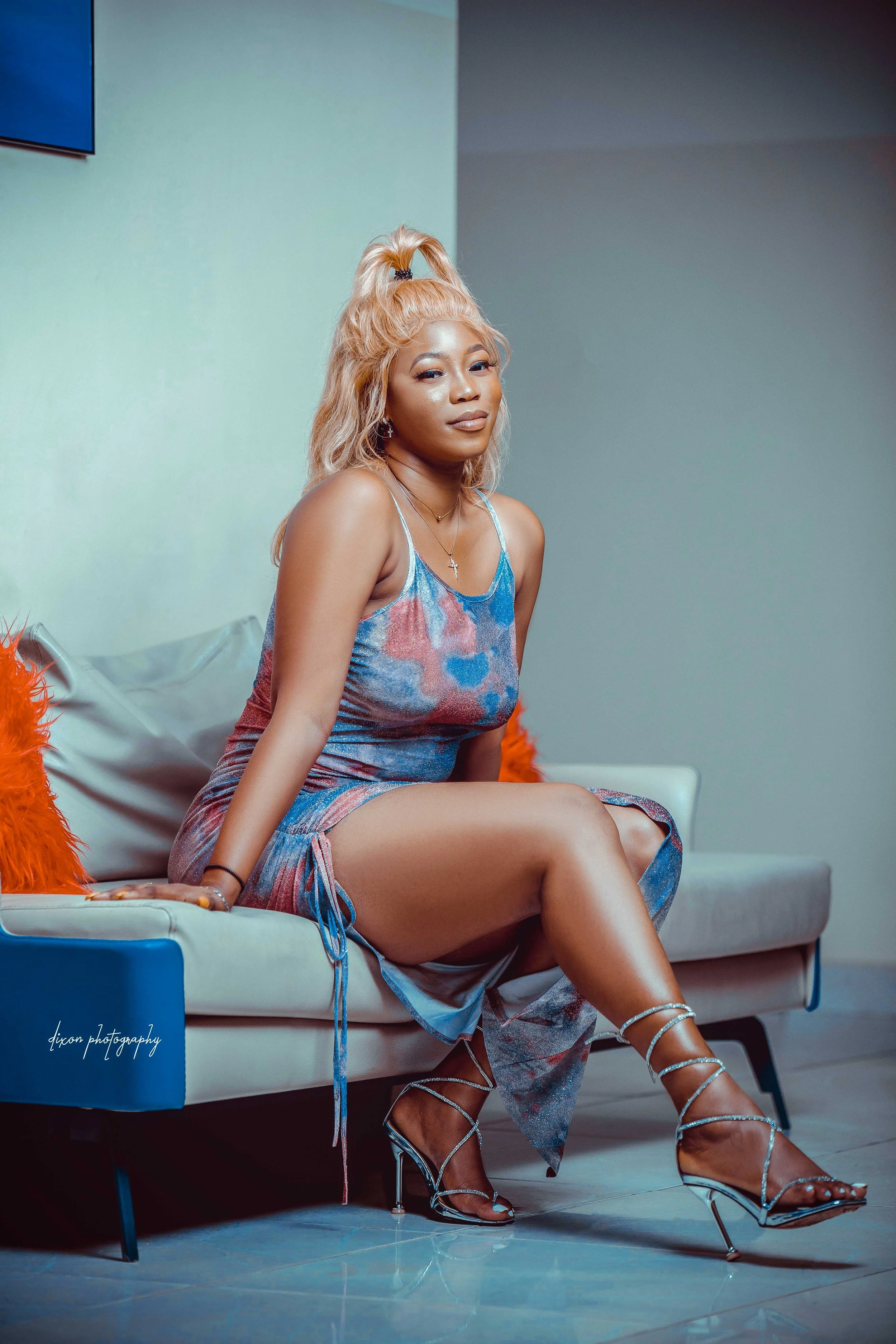 A woman with blonde hair styled in a high ponytail is sitting on a modern beige sofa with an orange cushion and a grey cushion, wearing a colorful tie-dye dress and shiny silver high-heeled sandals, posing indoors.