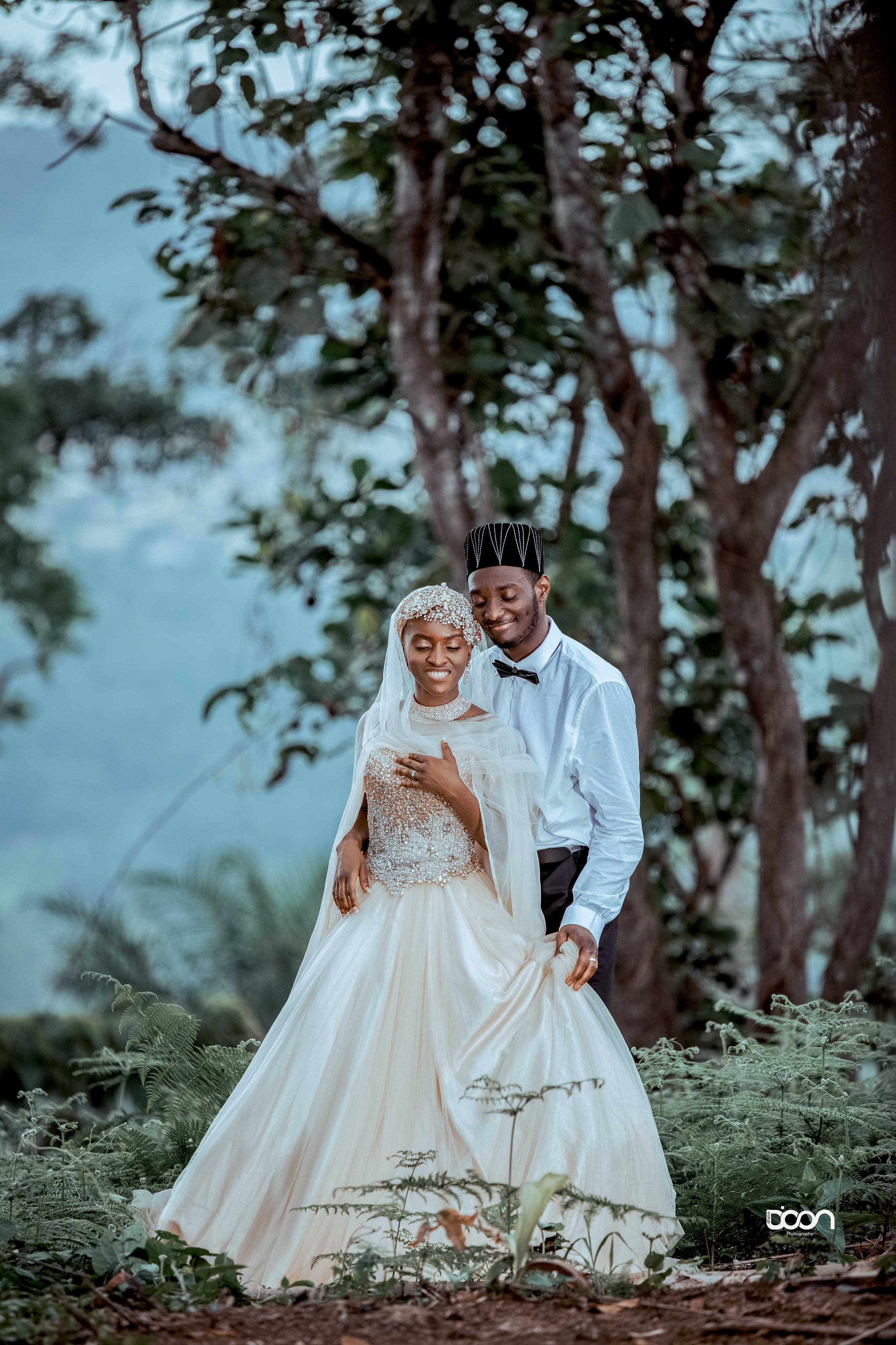 A bride and groom in wedding attire standing outdoors among trees, smiling, with the bride holding her gown and the groom holding her hand.