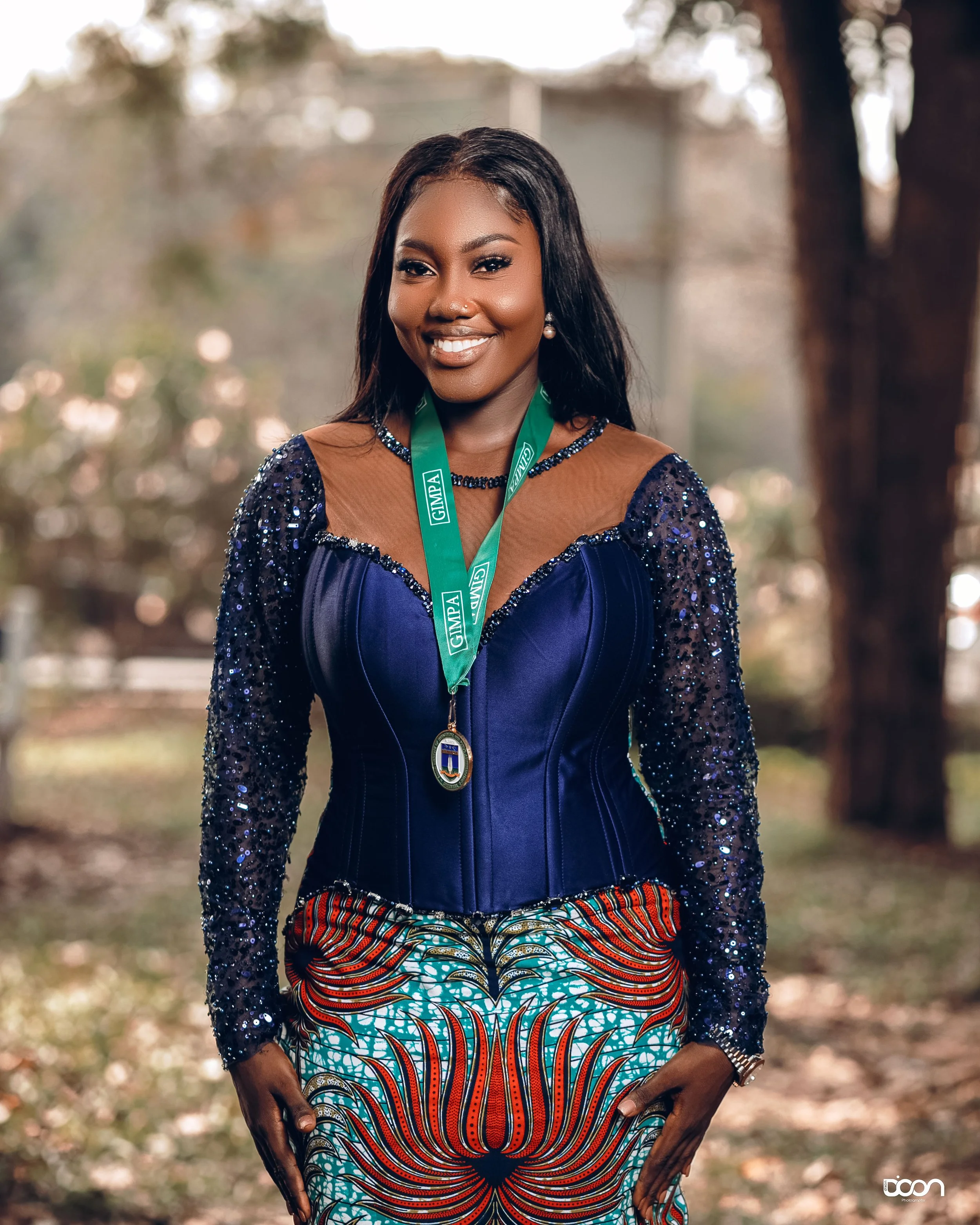 A woman wearing a navy blue corset top with beaded long sleeves, a patterned skirt with orange, black, turquoise, and white designs, and a green lanyard with a medal, standing outdoors. She has long black hair, pearl earrings, and is smiling.