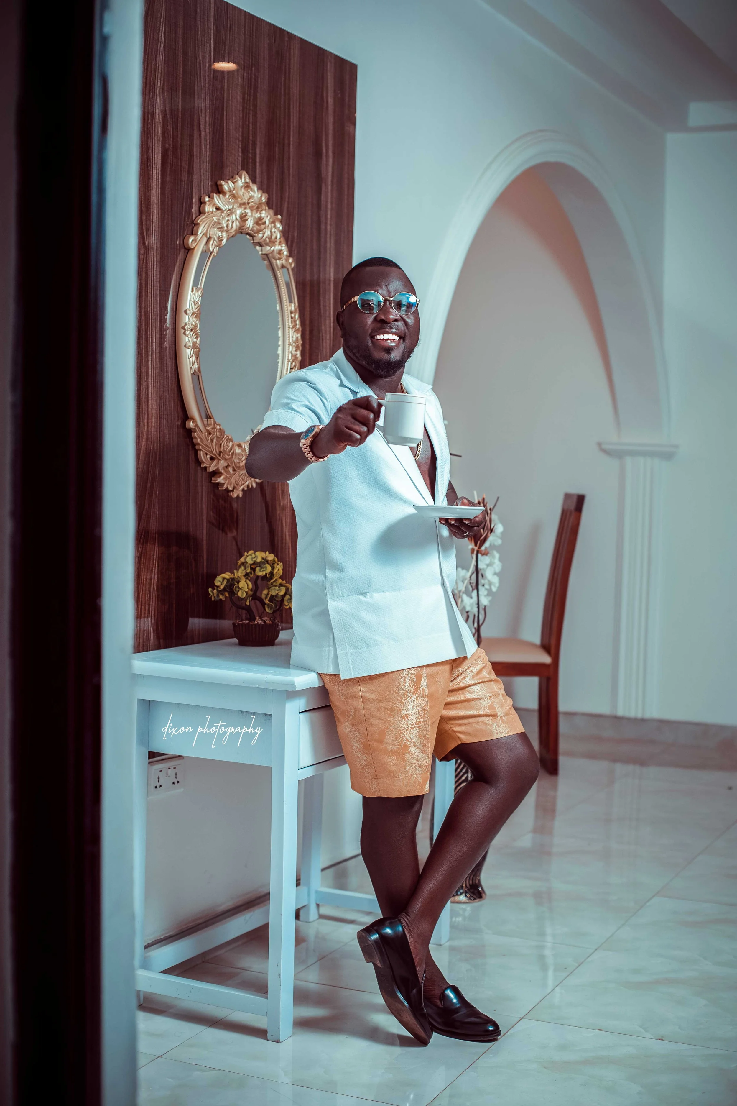 A man standing indoors, holding a coffee cup and a saucer, smiling, wearing glasses, a white blazer, beige shorts, black shoes, and a watch, with a decorative mirror, flower arrangements, and chairs in the background.
