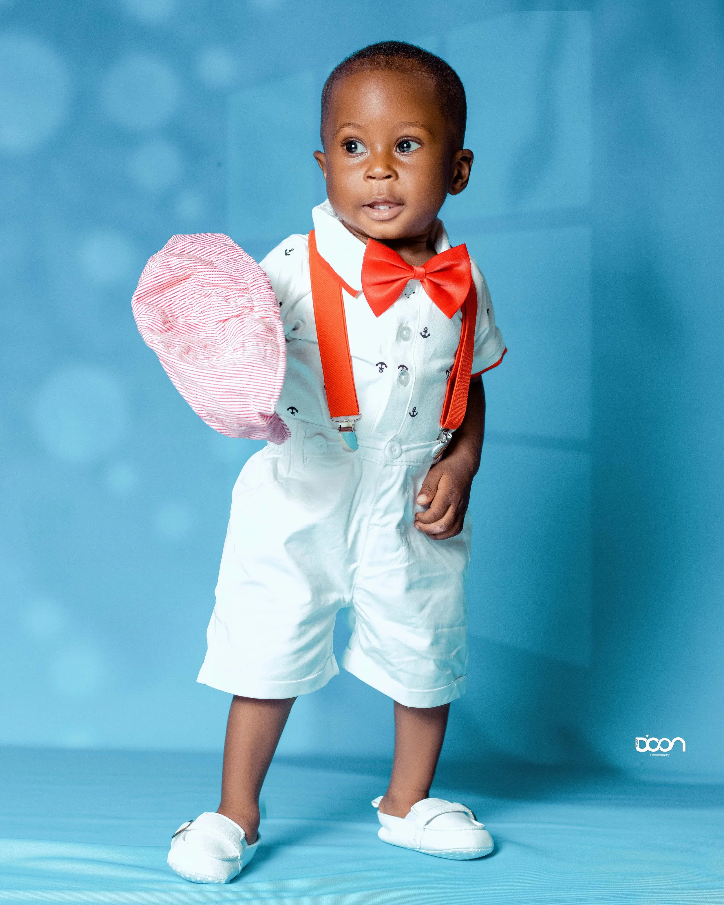 A young boy dressed in a white shirt with small anchor patterns, white shorts, red suspenders, and a red bow tie, standing against a blue background, holding a red and white striped hat in one hand.