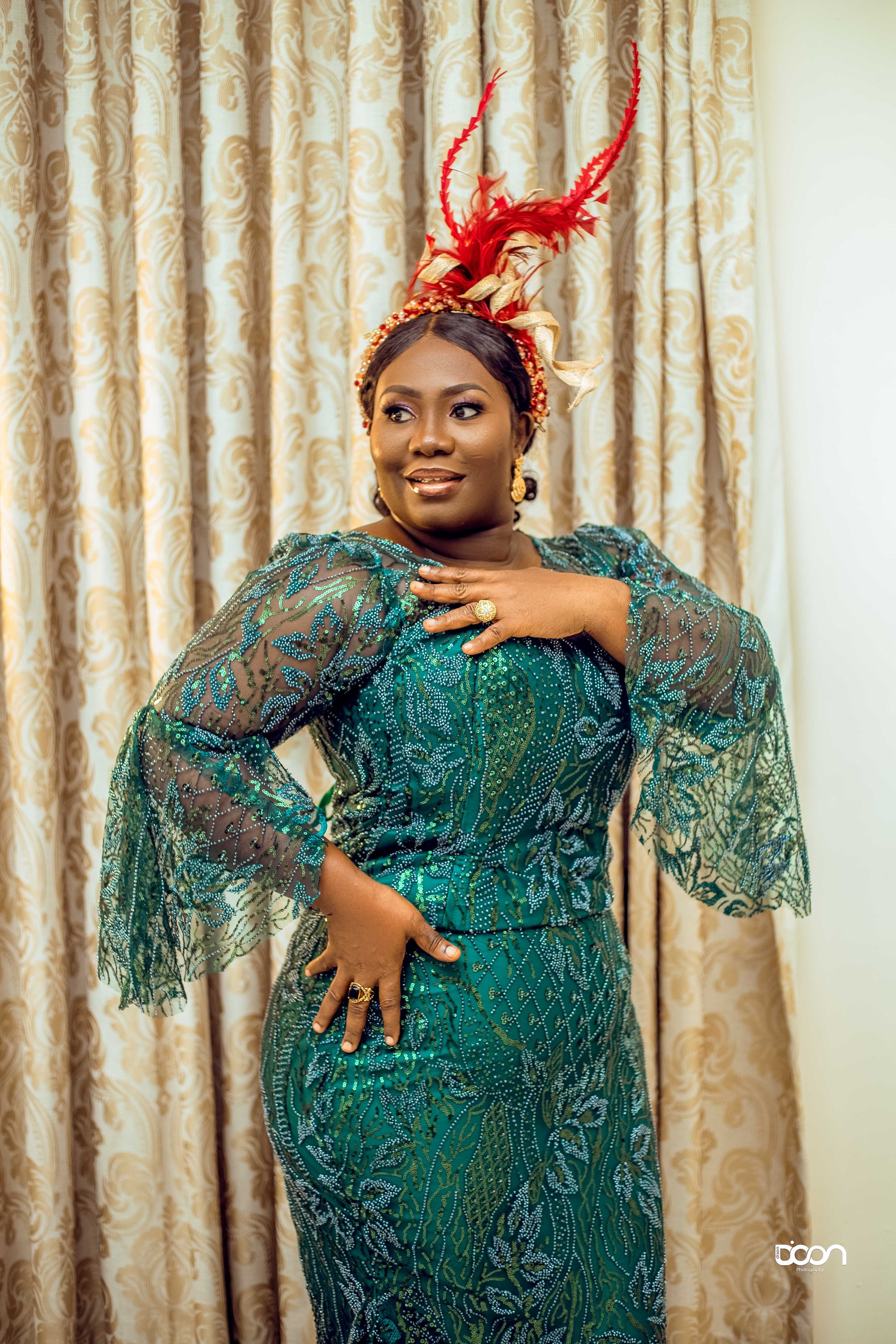 A woman in a vibrant green and blue traditional dress with intricate embroidery and sheer, ruffled sleeves, poses in front of patterned curtains, wearing a red feathered headpiece and gold jewelry.