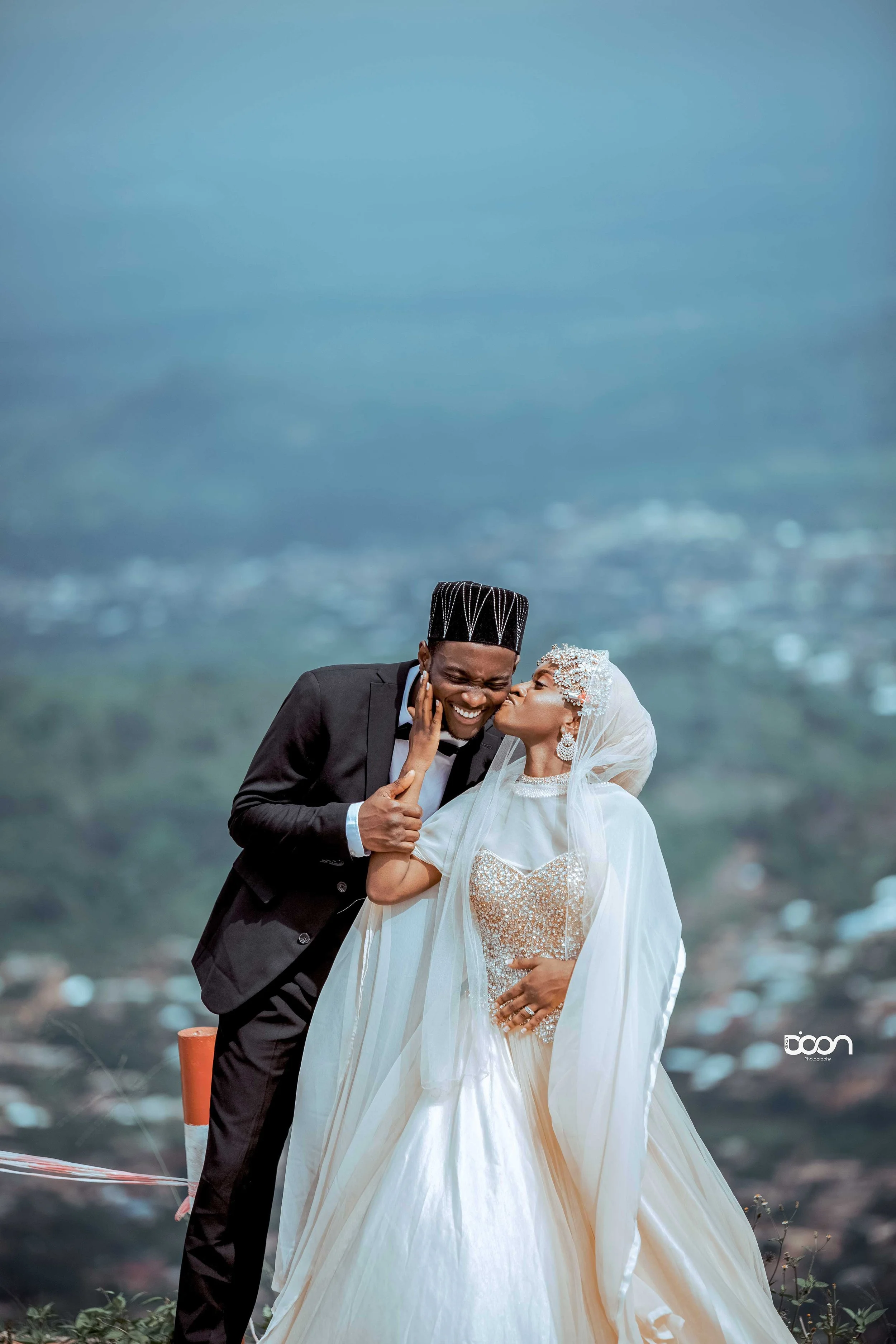 A bride and groom on their wedding day, with a scenic background. The groom is wearing a black suit and tie, smiling as he leans towards the bride. The bride is wearing a white gown with intricate embellishments, a veil, and matching jewelry, giving 