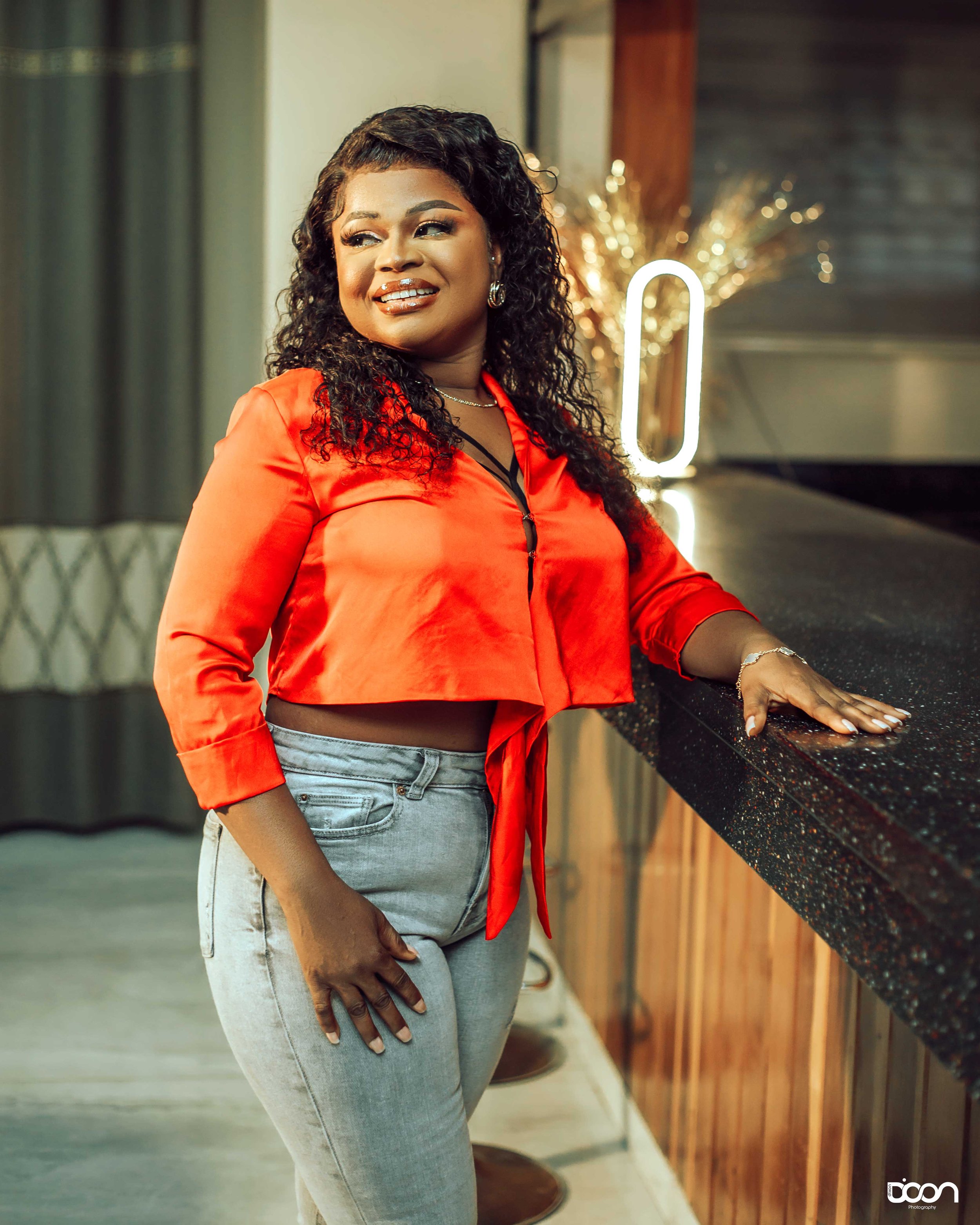 A woman with curly black hair and wearing a red satin blazer and light gray jeans standing at a bar counter, smiling and looking to the side.