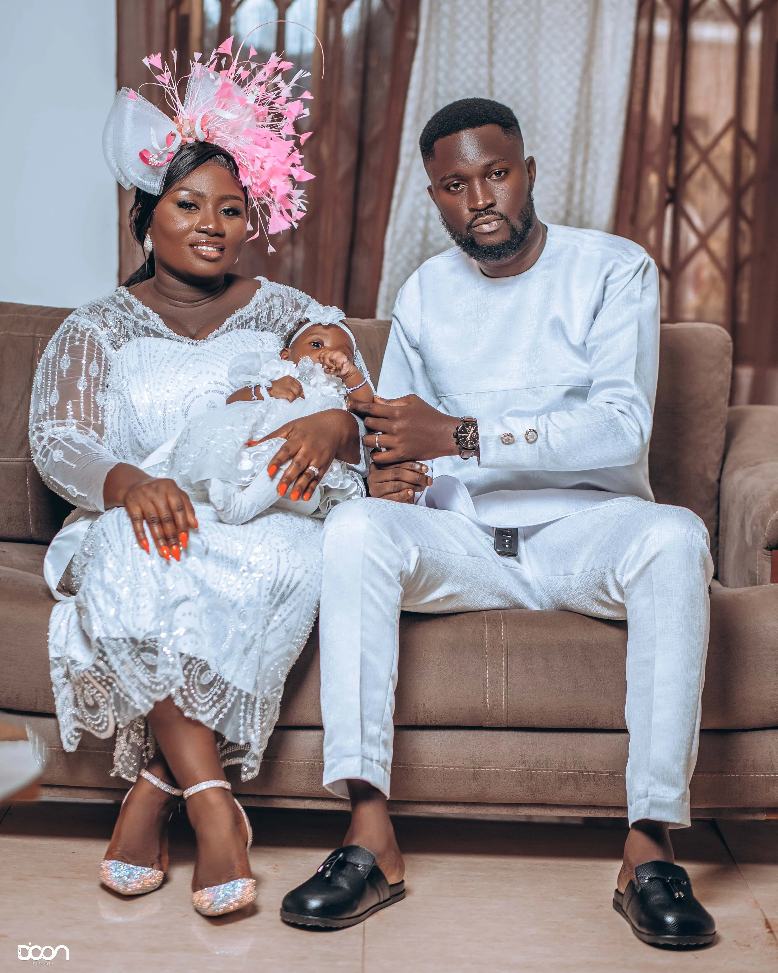 A couple sitting on a sofa with a baby dressed in white, the woman wearing a white lace dress and a pink elaborate hat, and the man in a white outfit, with the woman holding the baby.