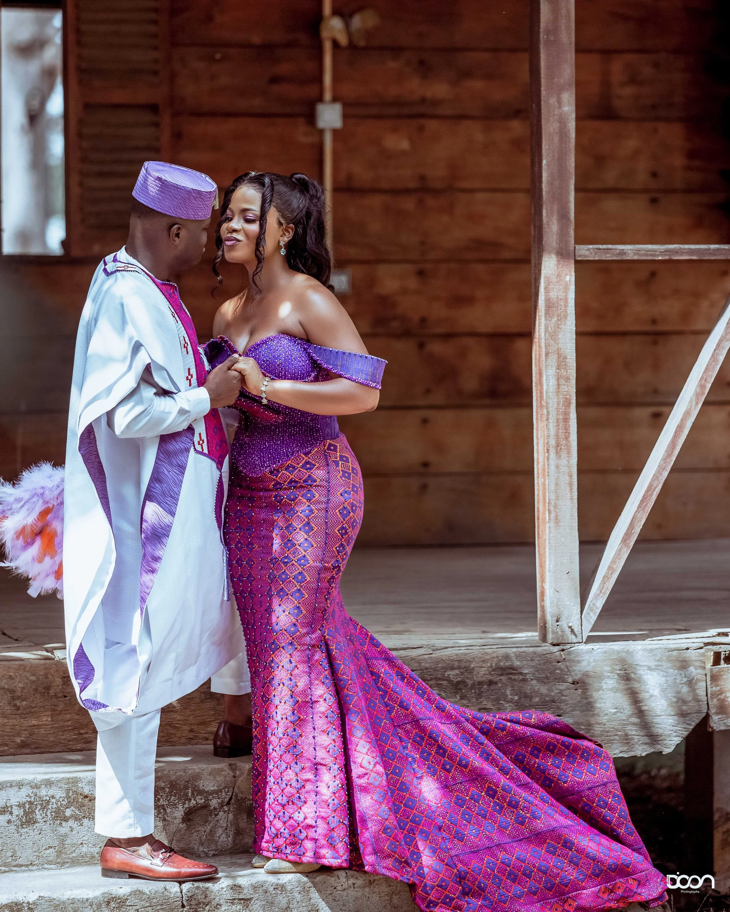 A couple dressed in traditional Nigerian attire standing on steps inside a wooden structure, holding hands and looking at each other.