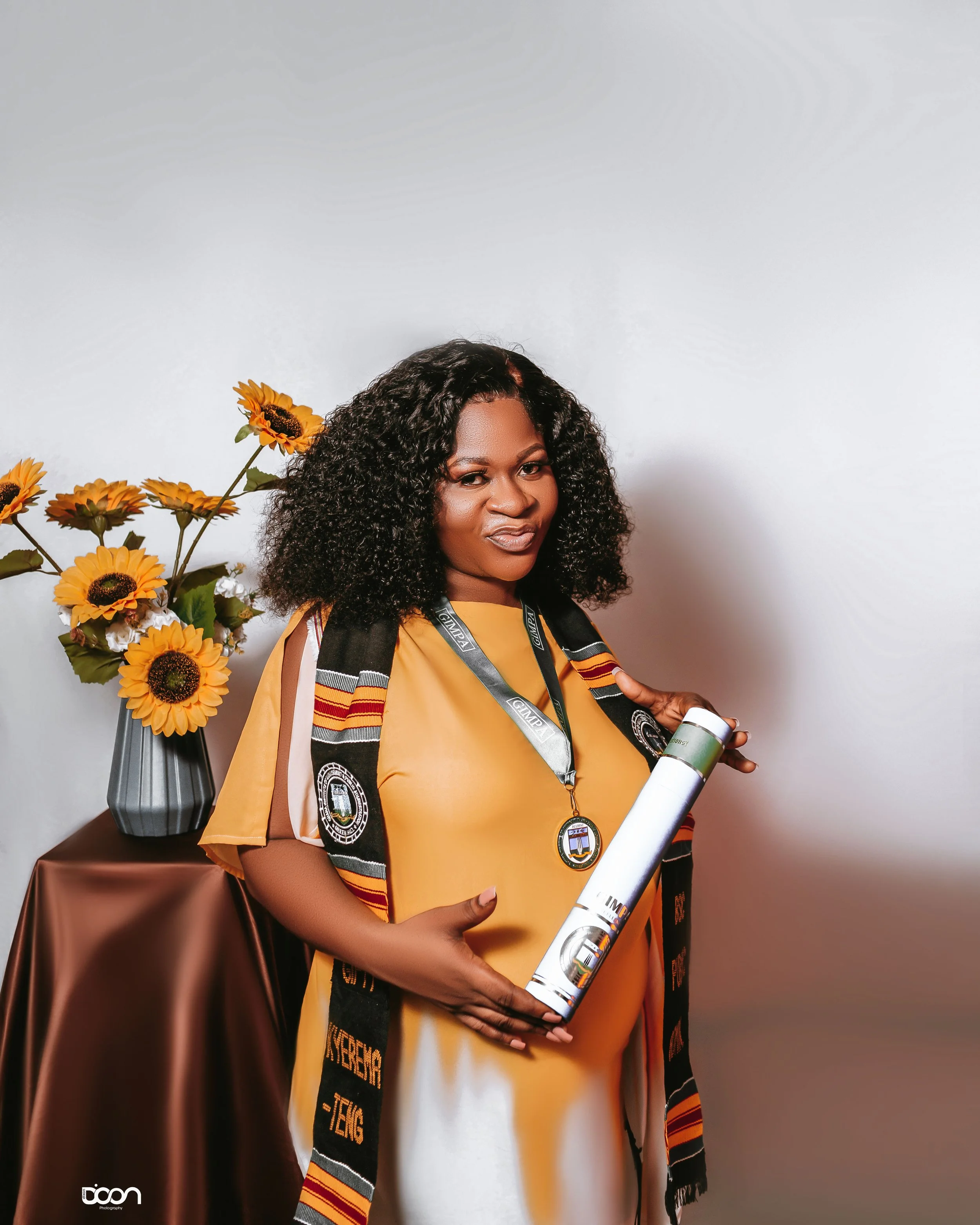 A woman in academic regalia holding a rolled-up document, standing beside a table with a bouquet of sunflowers in a vase.