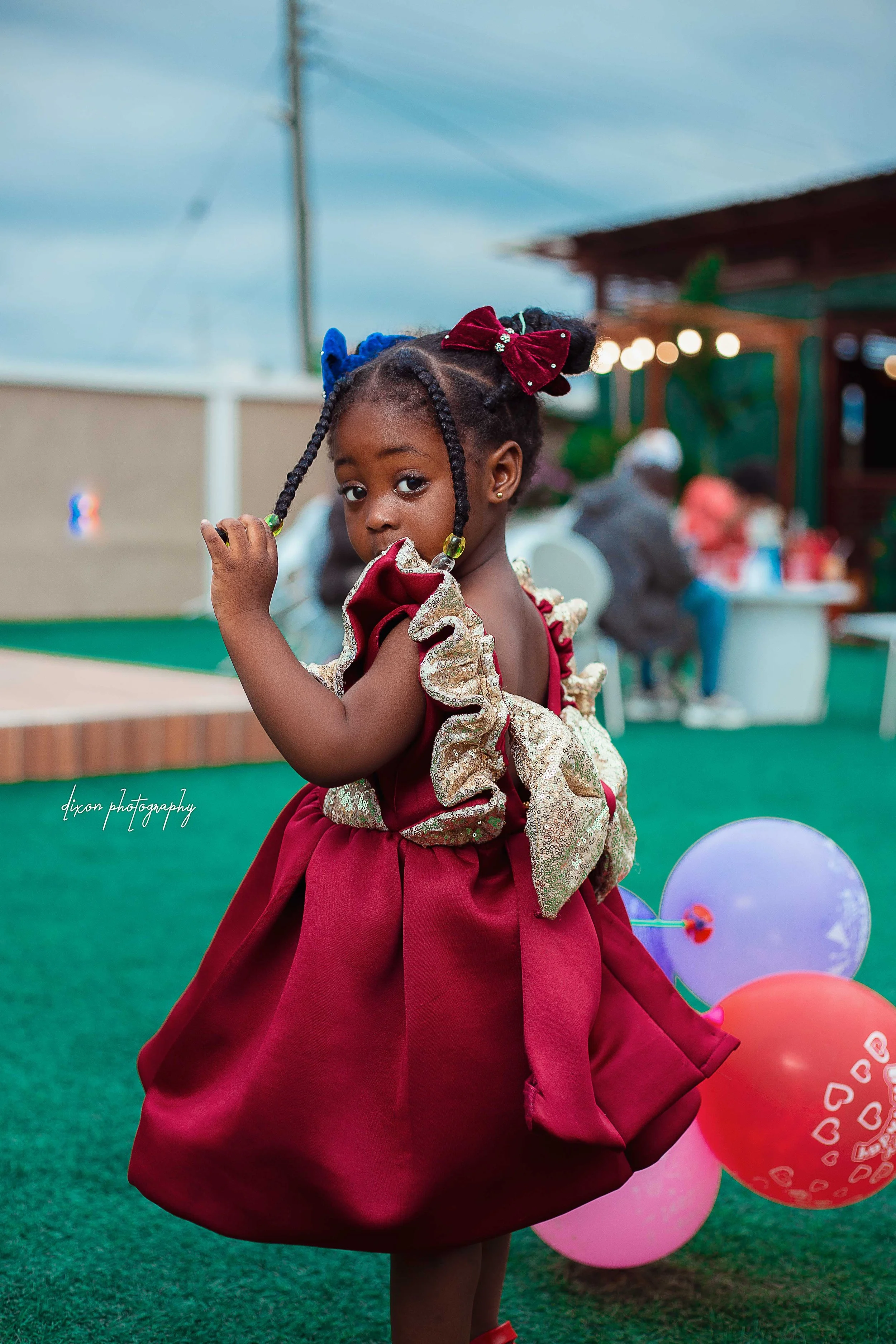 A young girl with braided hair adorned with colorful bows, wearing a fancy red and gold dress, standing on a green outdoor surface at a celebration or party, with balloons in the background.