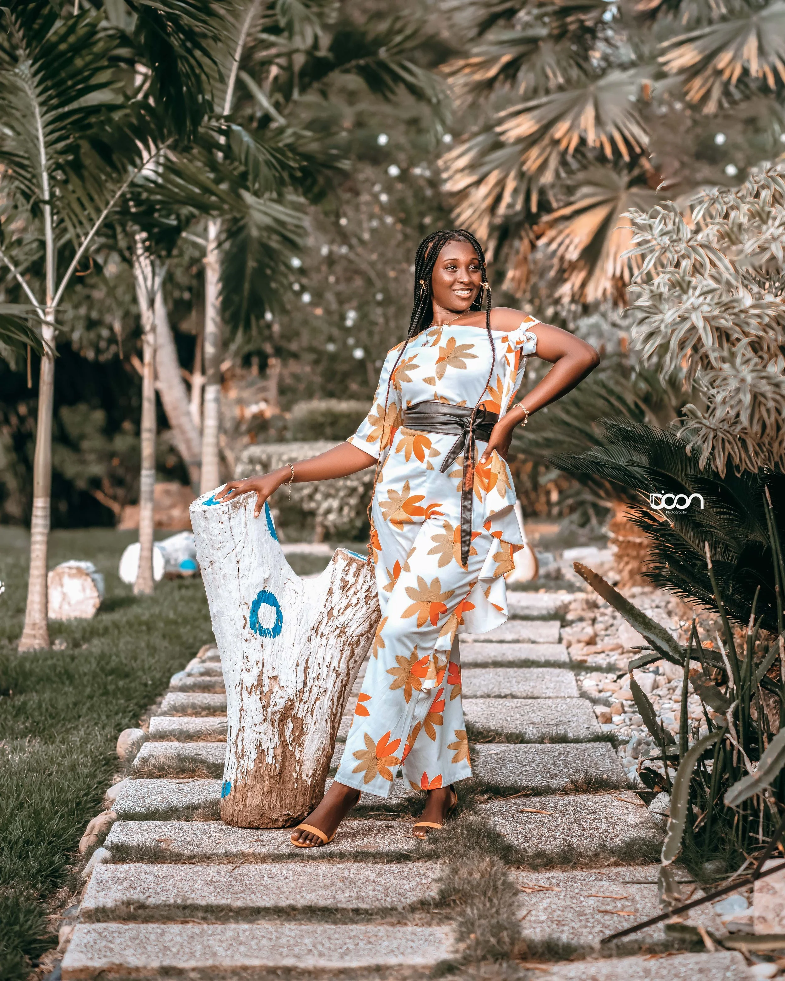 A woman in a floral patterned dress posing outdoors on a stone path, surrounded by tropical plants and trees.