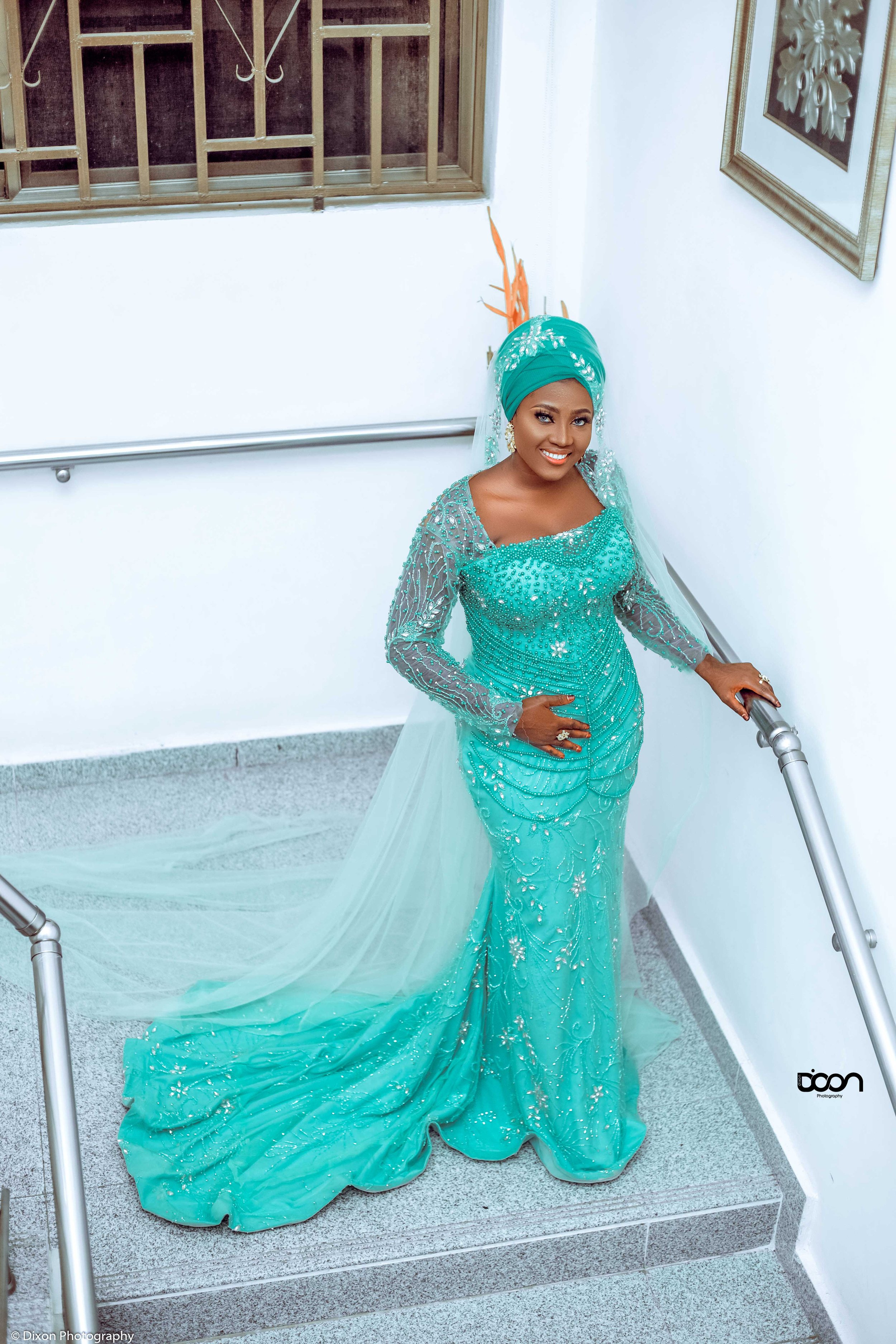 A woman dressed in an elegant turquoise gown with intricate beadwork, standing on a staircase with a smile.