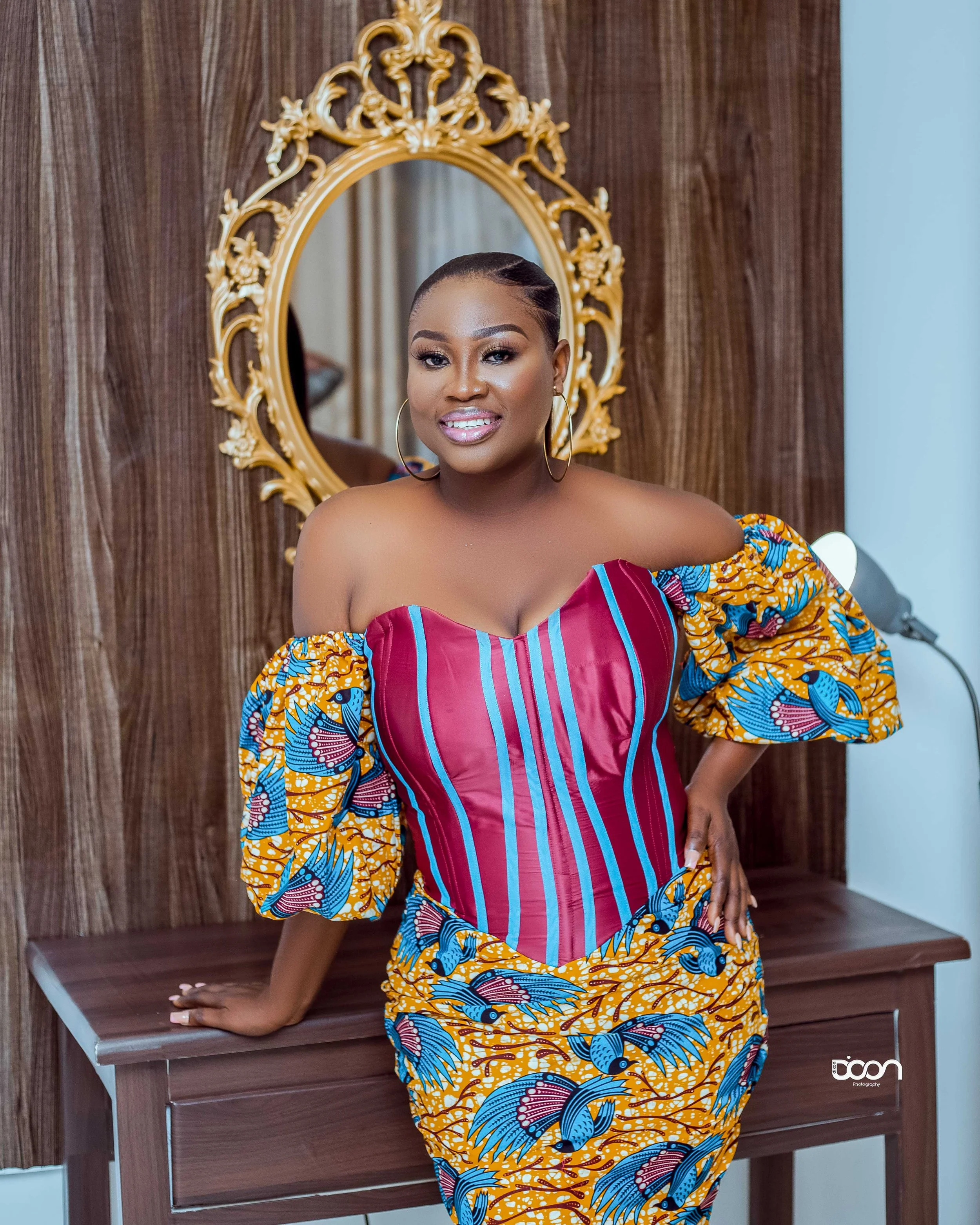 A woman in a colorful, off-the-shoulder dress standing in front of a wooden wall with an ornate gold mirror behind her.
