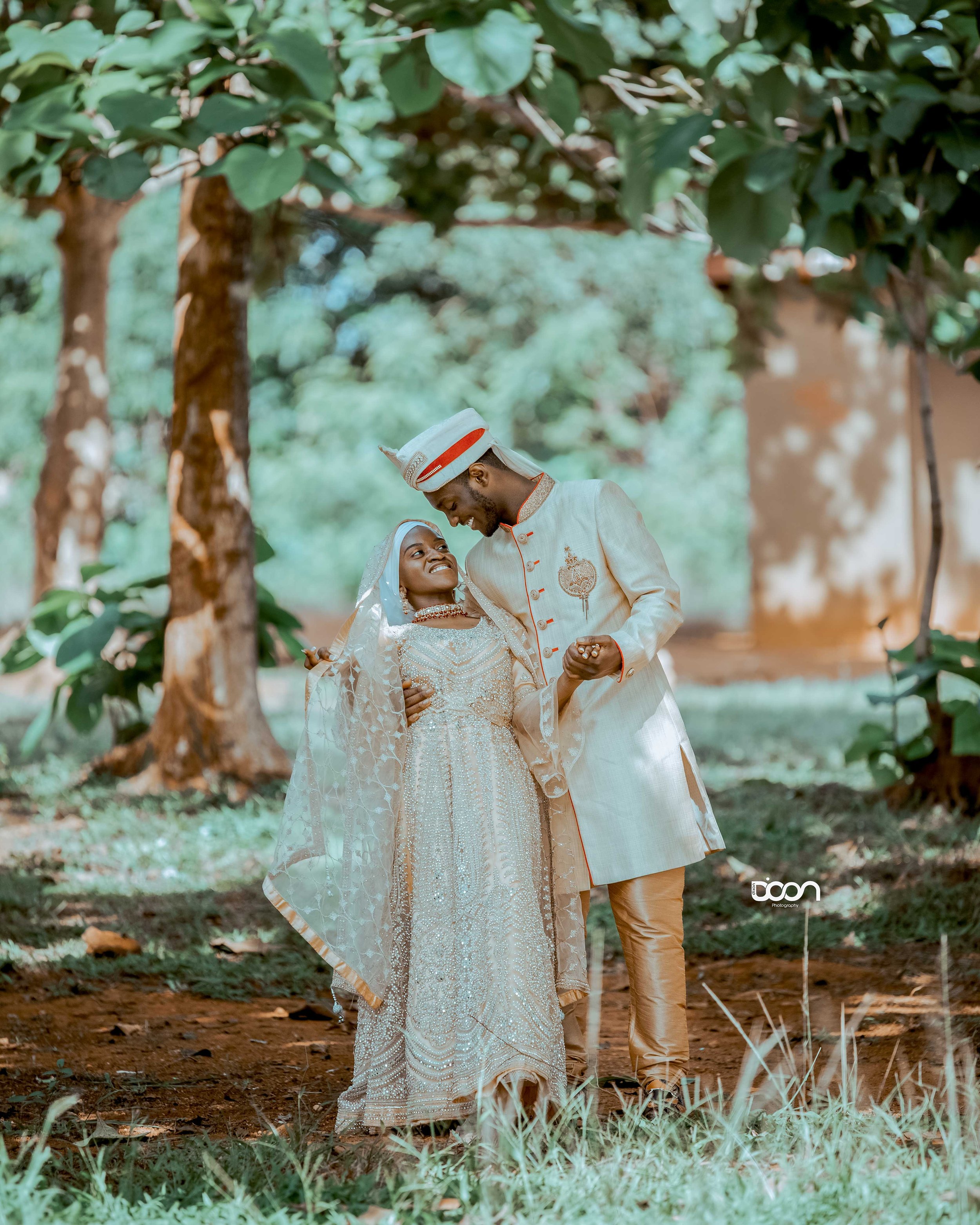 A couple dressed in traditional wedding attire dancing outdoors under trees, smiling at each other.